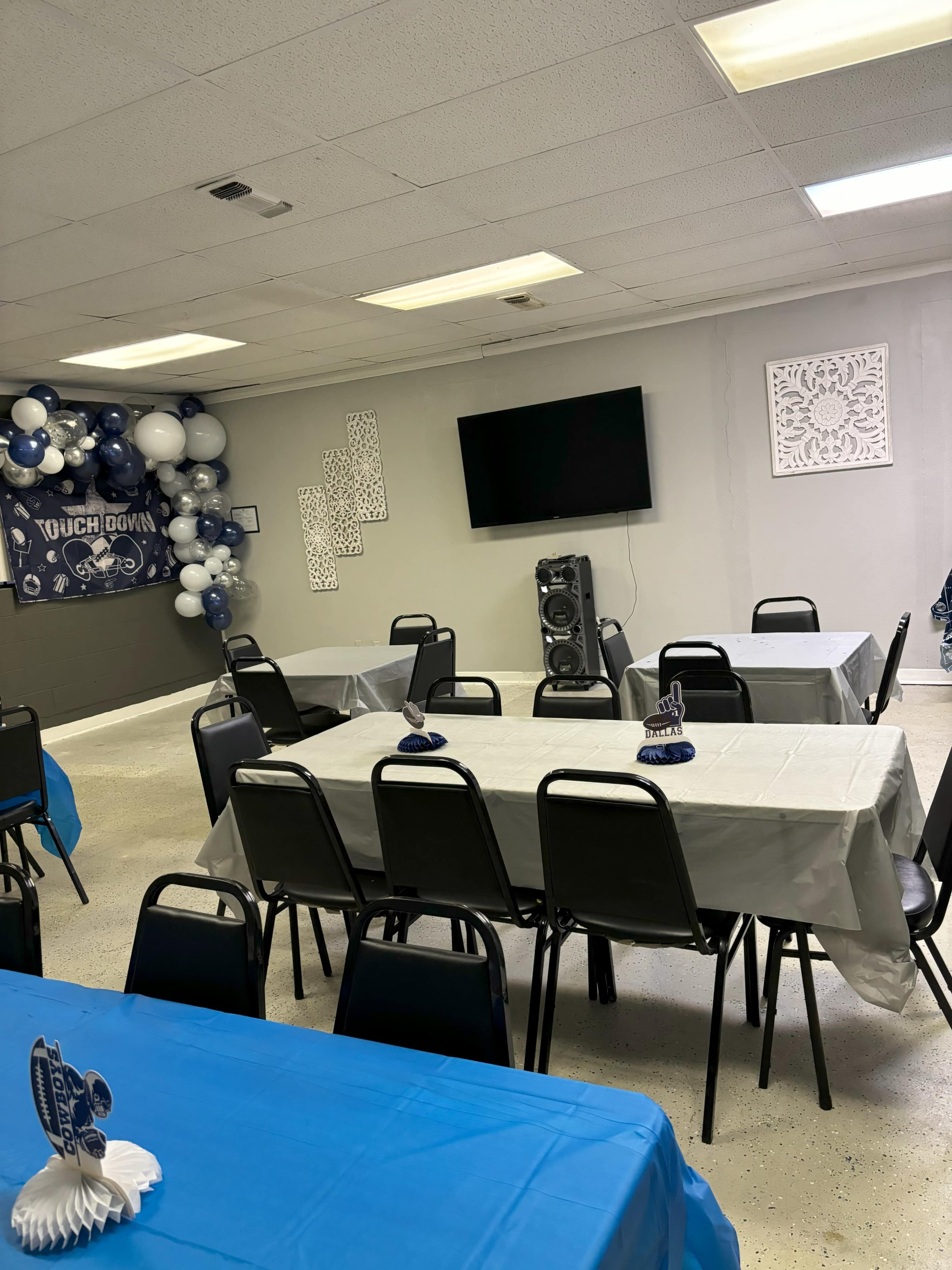 The image shows a room set up for an event, featuring several tables covered with tablecloths, a large television screen on the wall, and decorations of blue and white balloons.