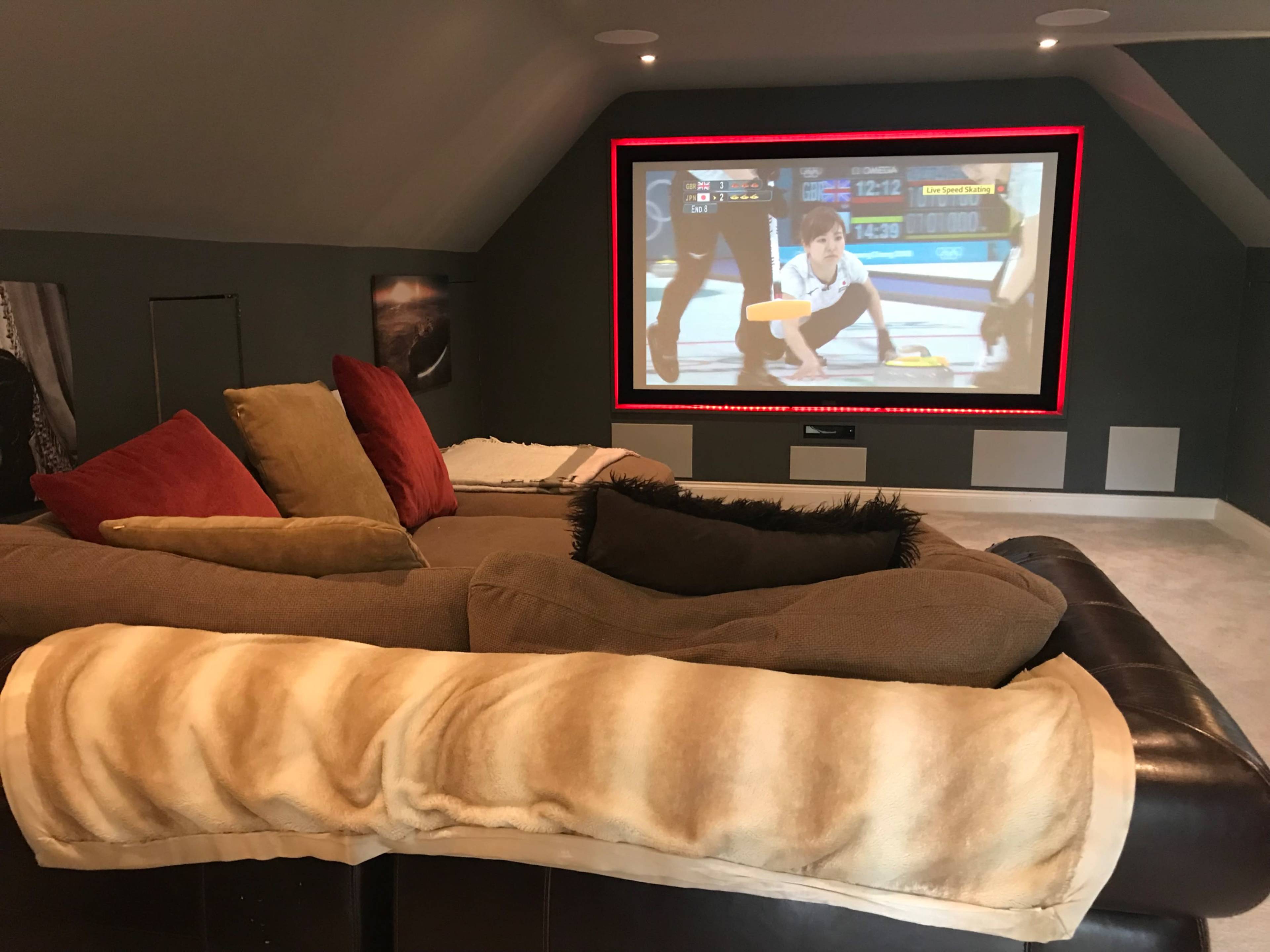 A home theater room features a large screen displaying an event, with a sectional sofa covered in blankets and cushions in the foreground.