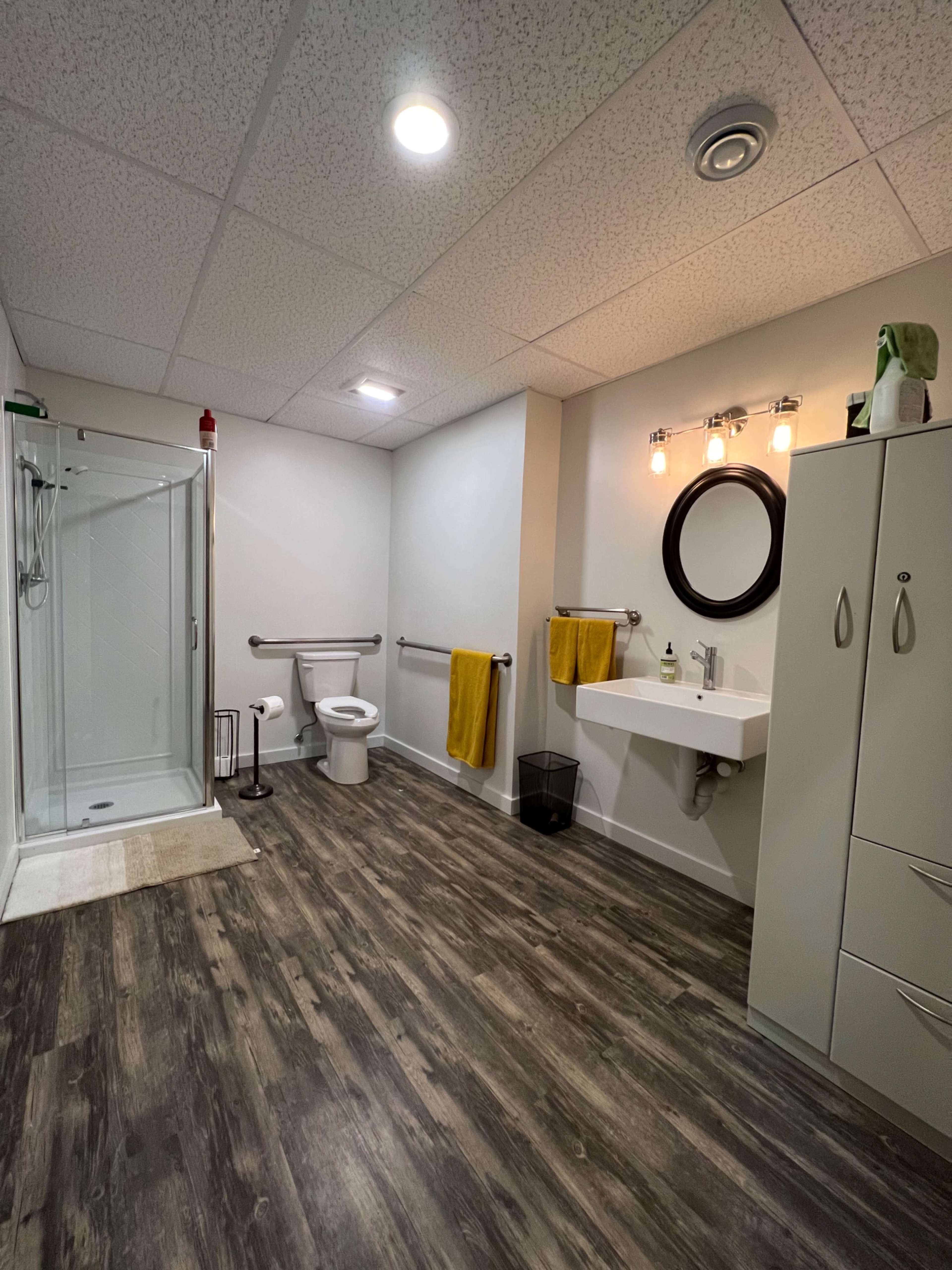 A modern bathroom featuring a shower stall, toilet, and a wall-mounted sink with a round mirror, accented by two yellow towels and a cabinet.