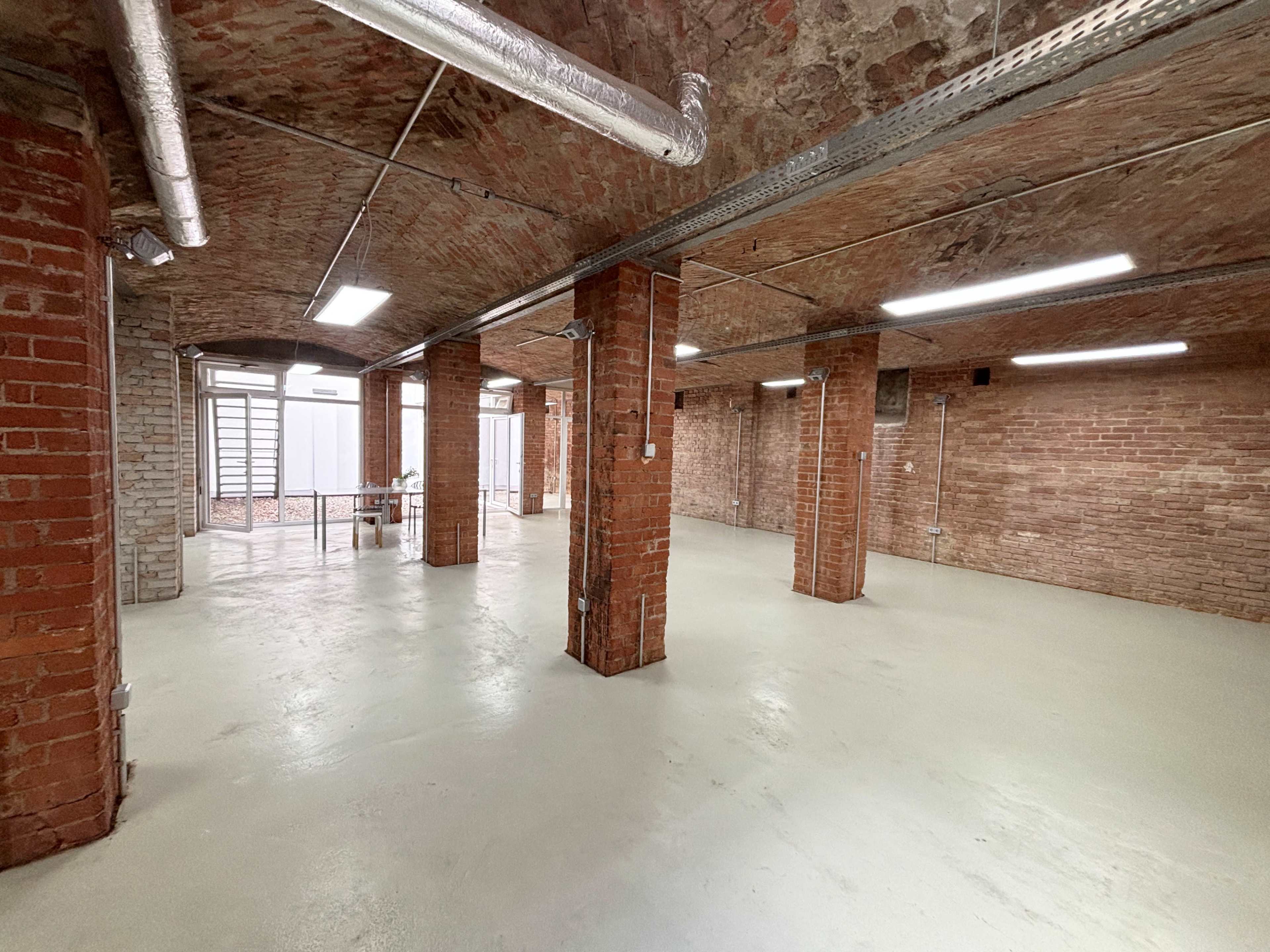 The image shows a spacious interior room with exposed brick walls, a concrete floor, and several structural columns.