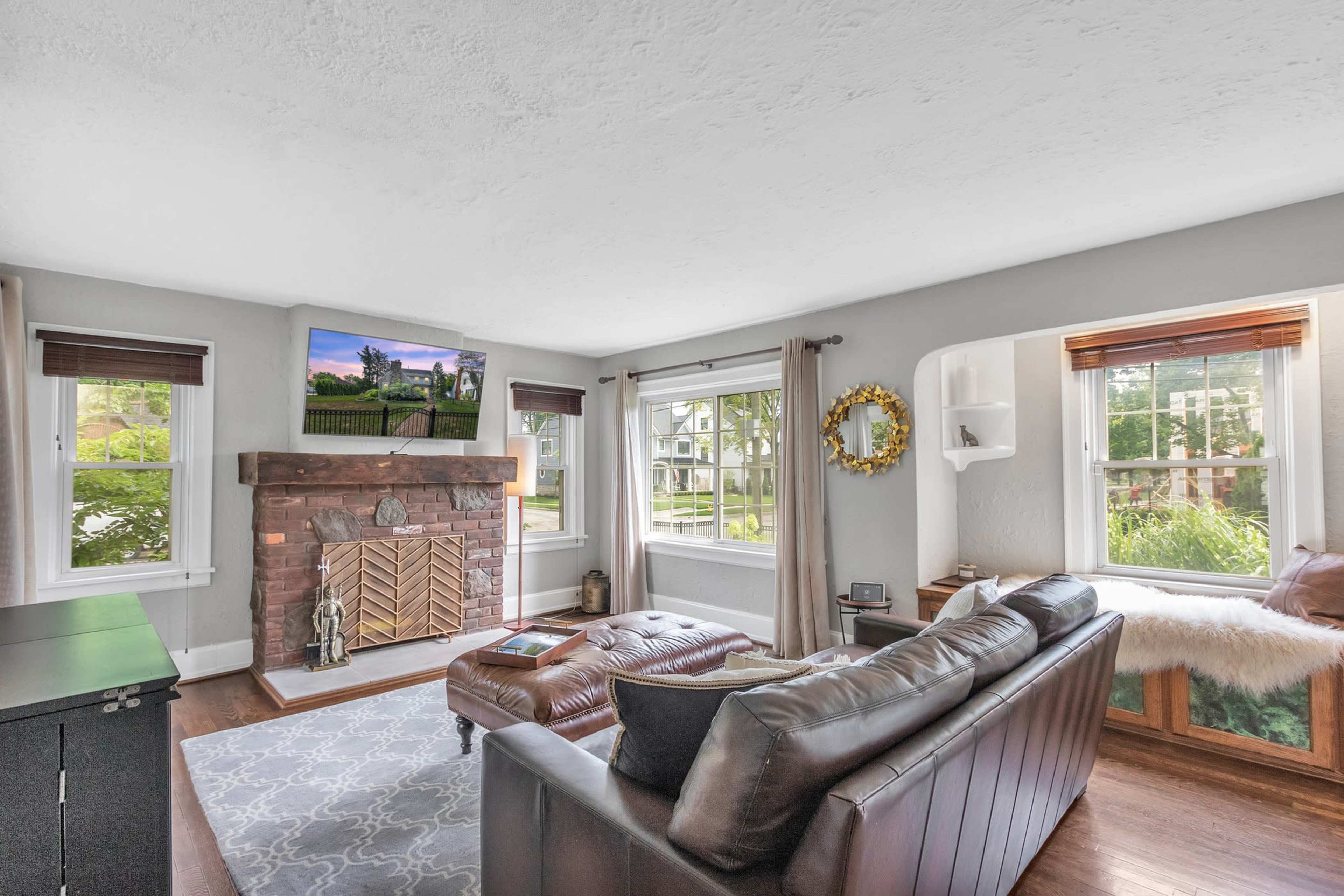 A living room features a brown leather sofa, a stone fireplace, and large windows with greenery visible outside.