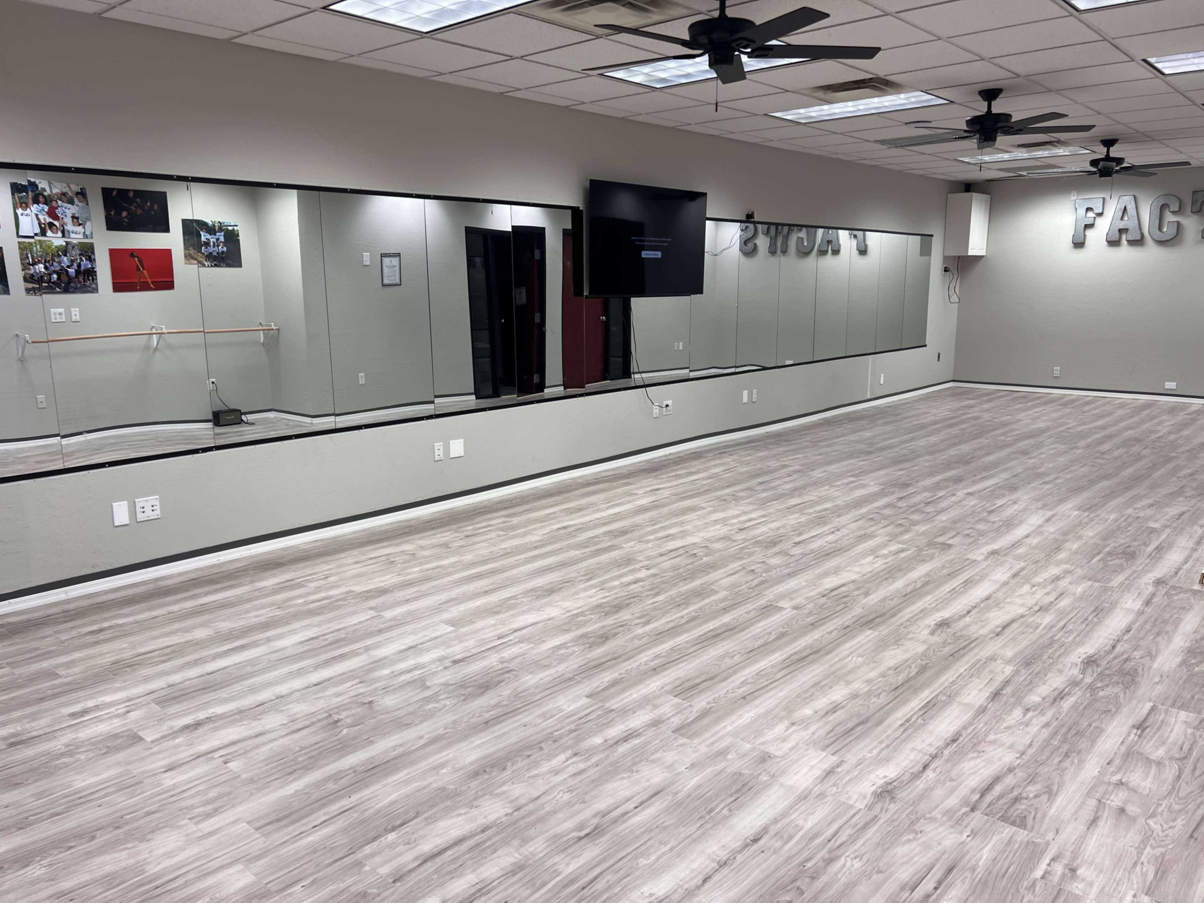 The image shows a dance studio with wooden flooring, mirrors on one wall, and a television mounted on another wall.