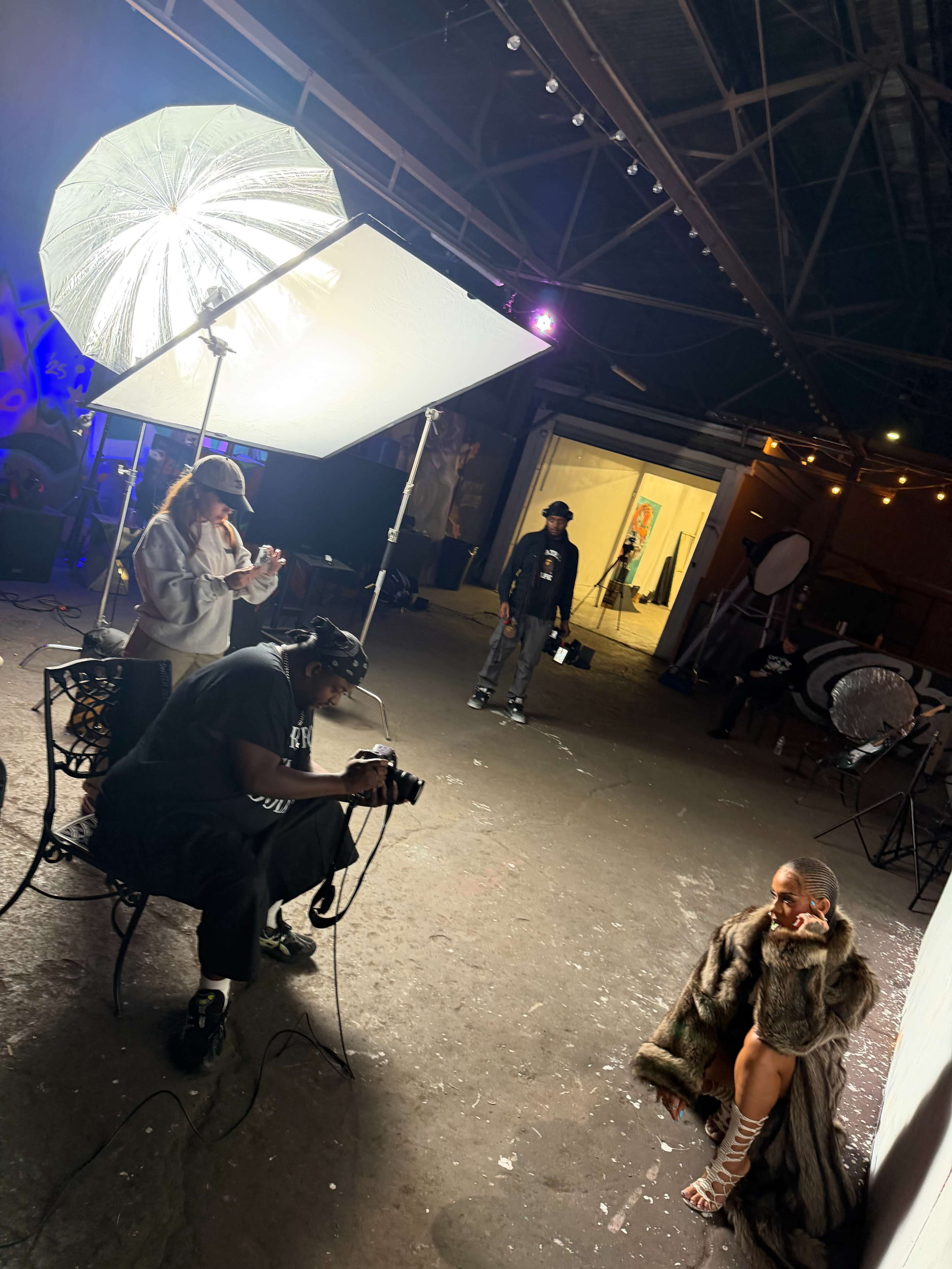 A photoshoot is taking place in an industrial space, with multiple people working around a model wearing a fur coat under bright lighting.