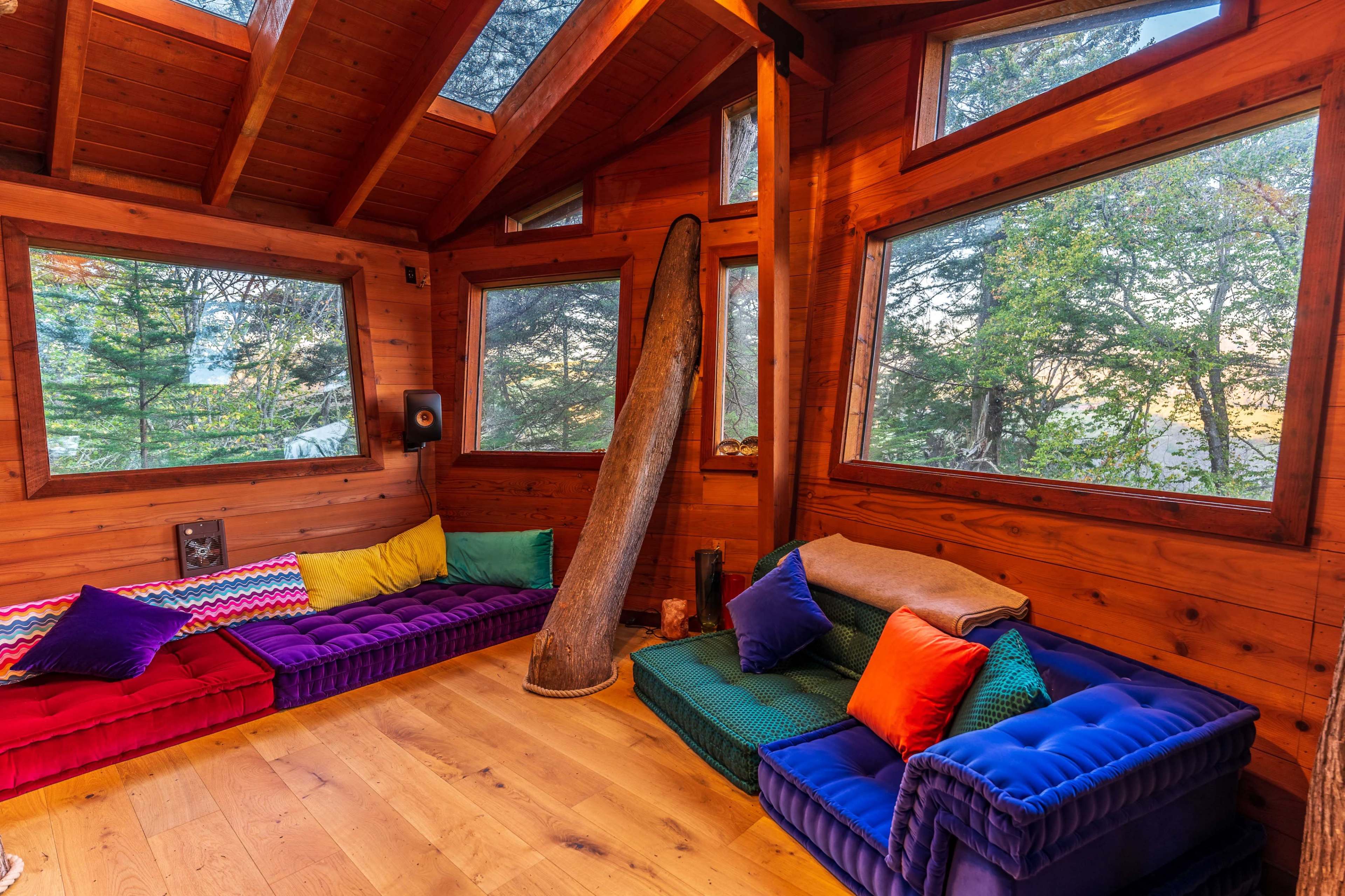 The interior of a wooden cabin features colorful couches with large windows overlooking a forested area.