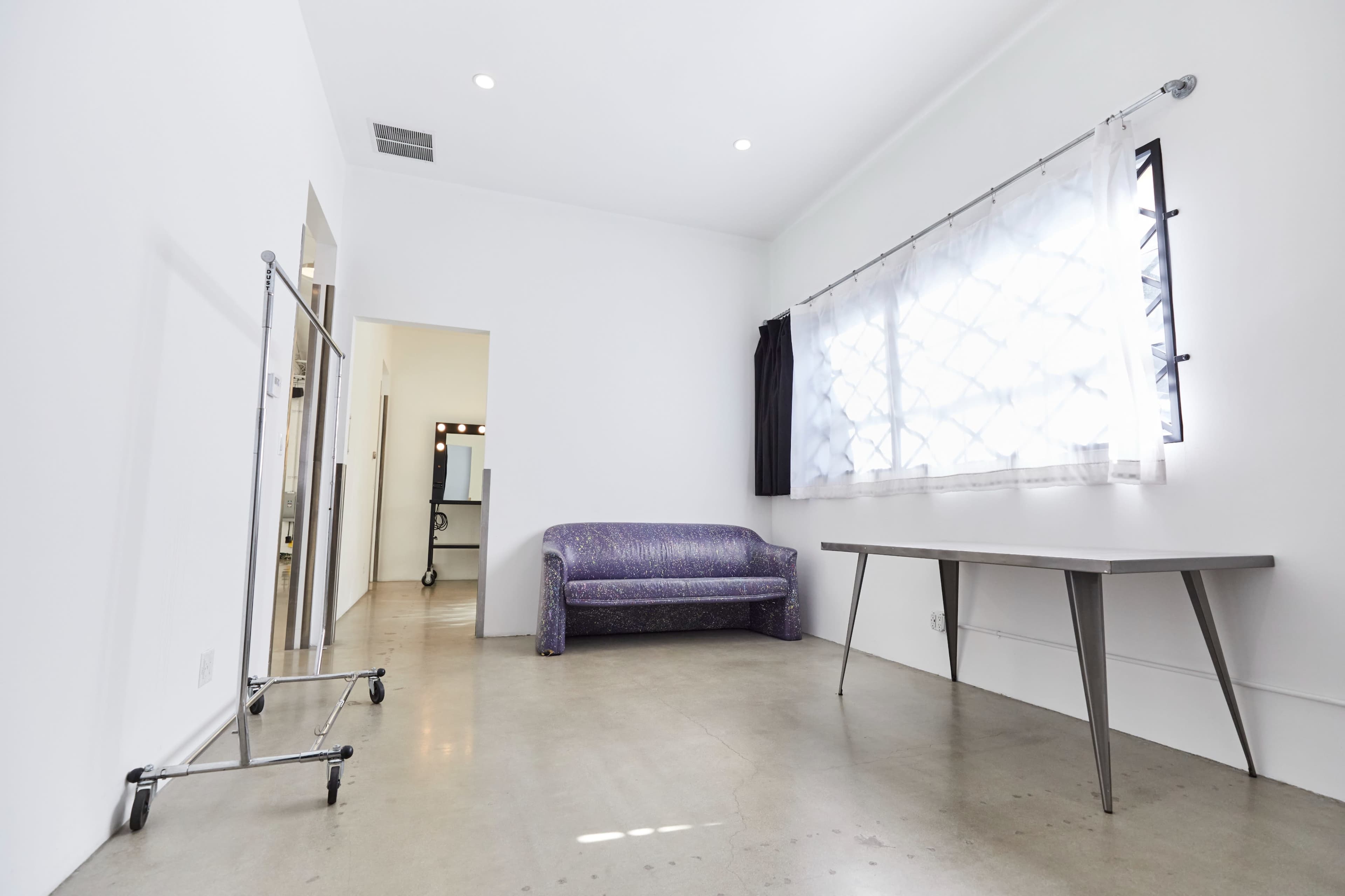 A sparsely furnished room features a purple couch, a table, and a clothing rack near a window with sheer curtains.