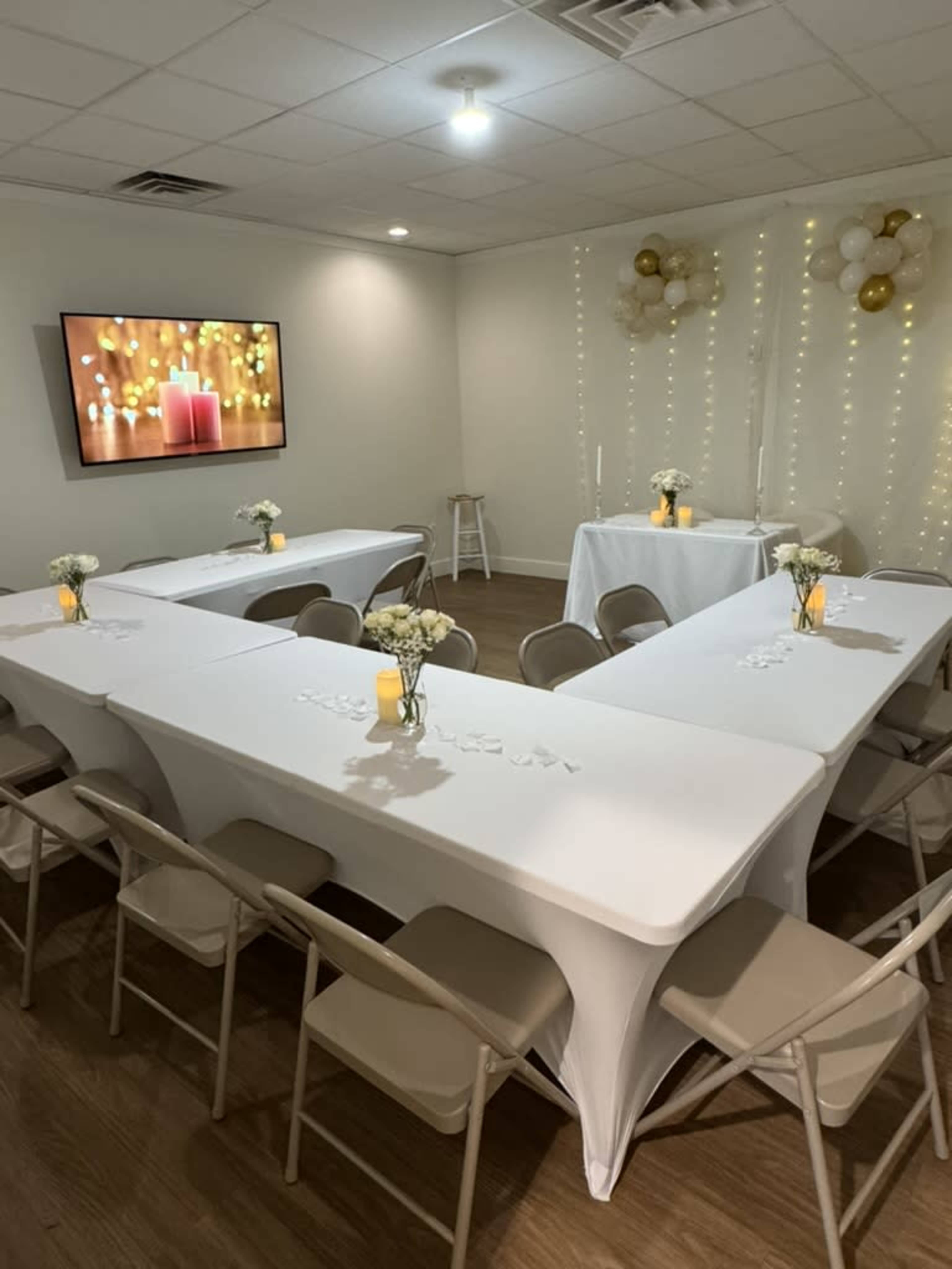 The image shows a decorated room with white tables arranged in a U-shape, featuring floral centerpieces and candles, with soft lighting and a screen displaying candles in the background.