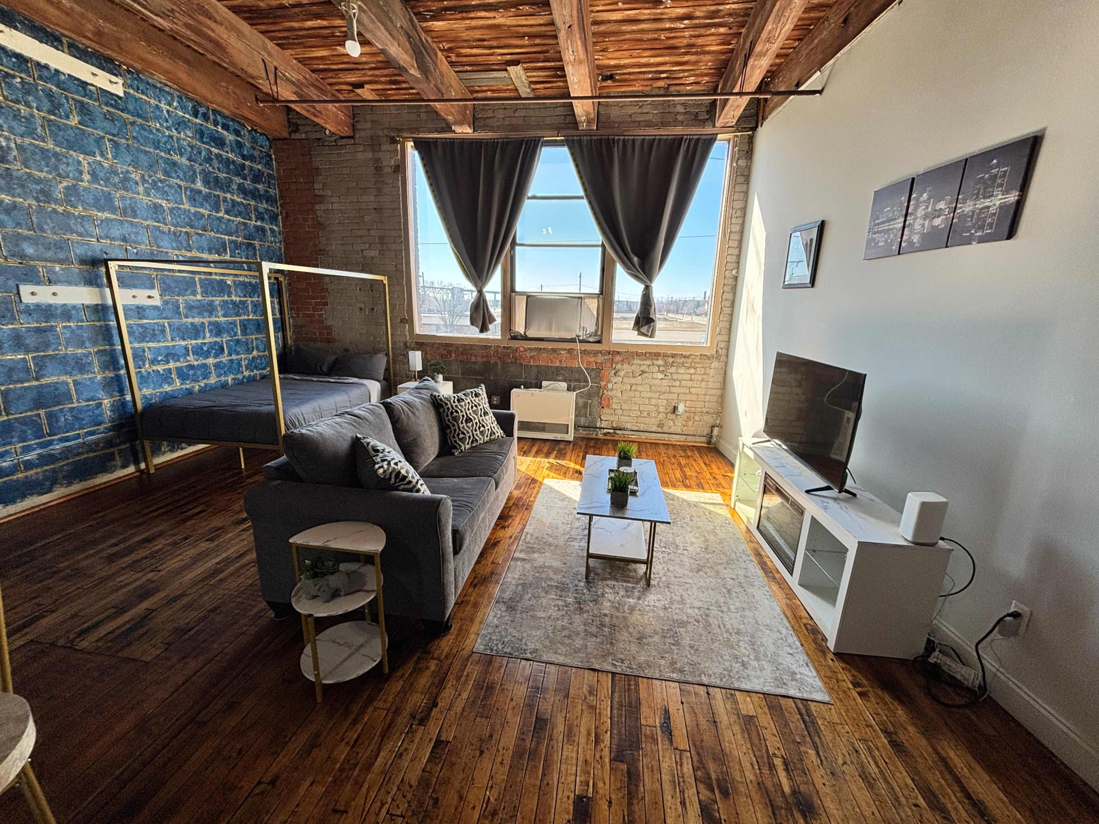The image shows a spacious, modern studio apartment featuring a living area with a gray sofa, a coffee table, and a television, alongside a bed area with a blue accent wall and large windows.