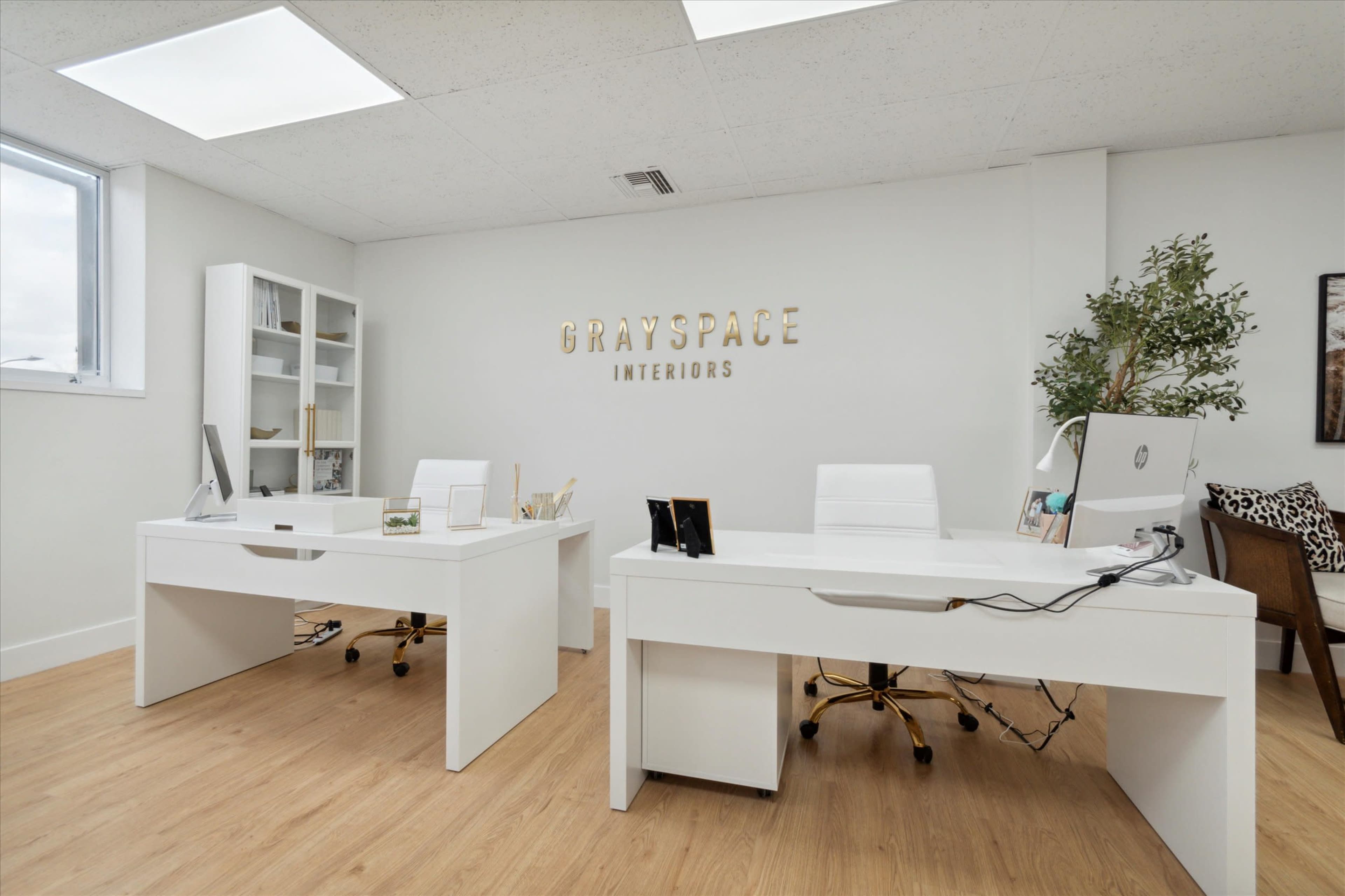 The image shows a modern office with two white desks, a plant, and a wall sign that reads "GRAYSPACE INTERIORS."