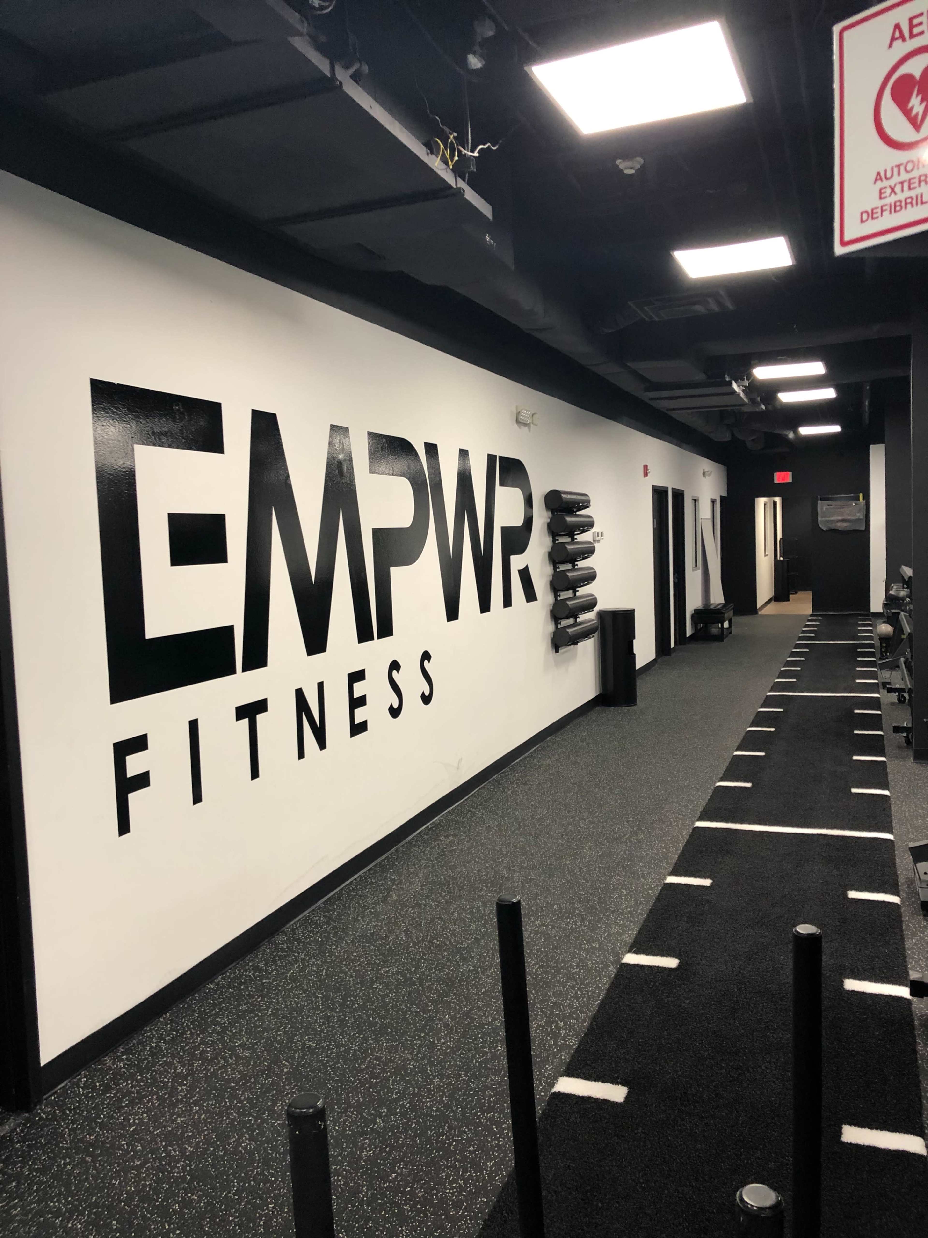The image shows a fitness facility hallway with a large "EMPWR FITNESS" sign on the wall and rubber flooring.