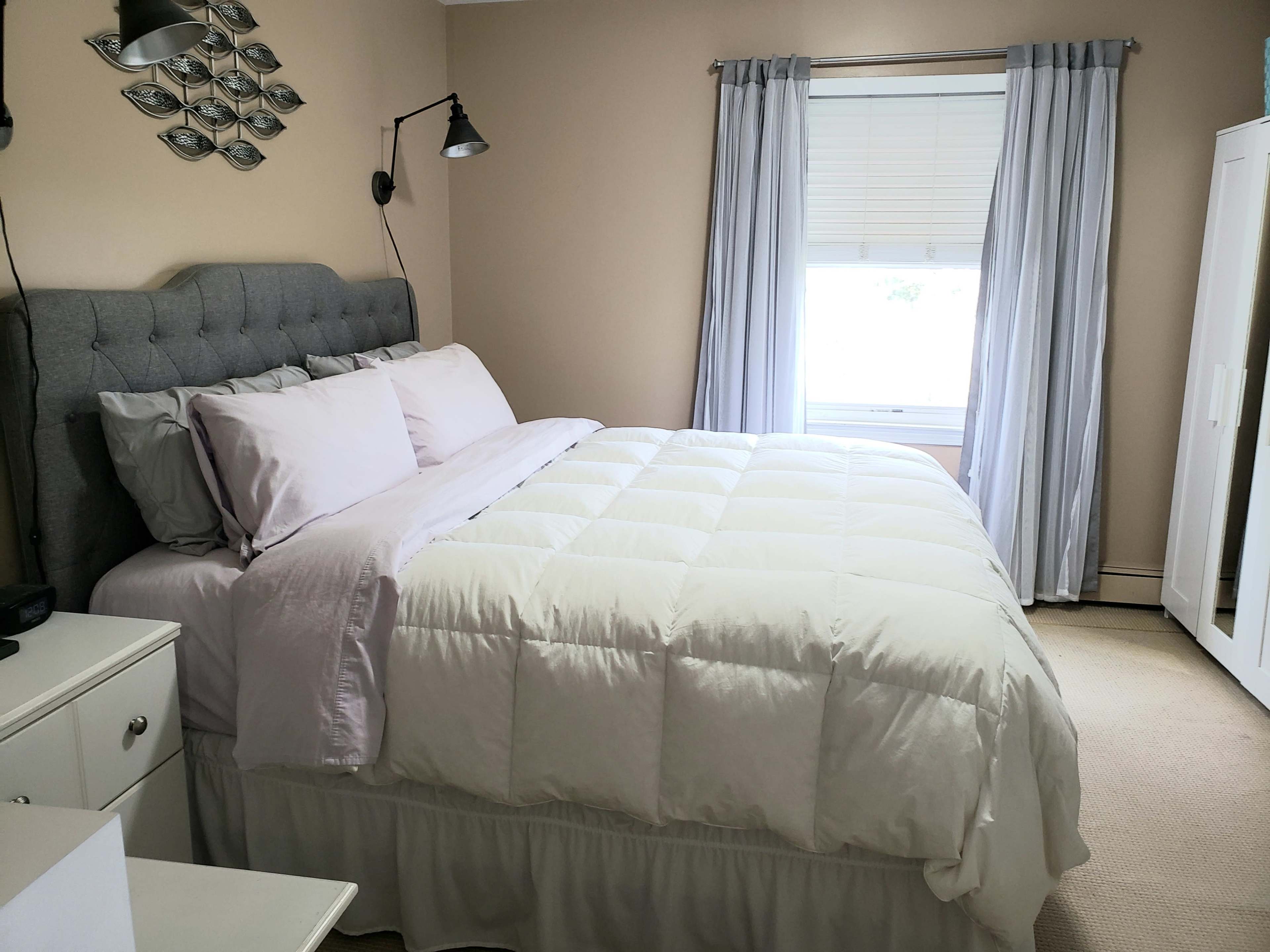 A neatly arranged bedroom with a gray upholstered headboard, a white comforter on the bed, and soft gray curtains over a window.