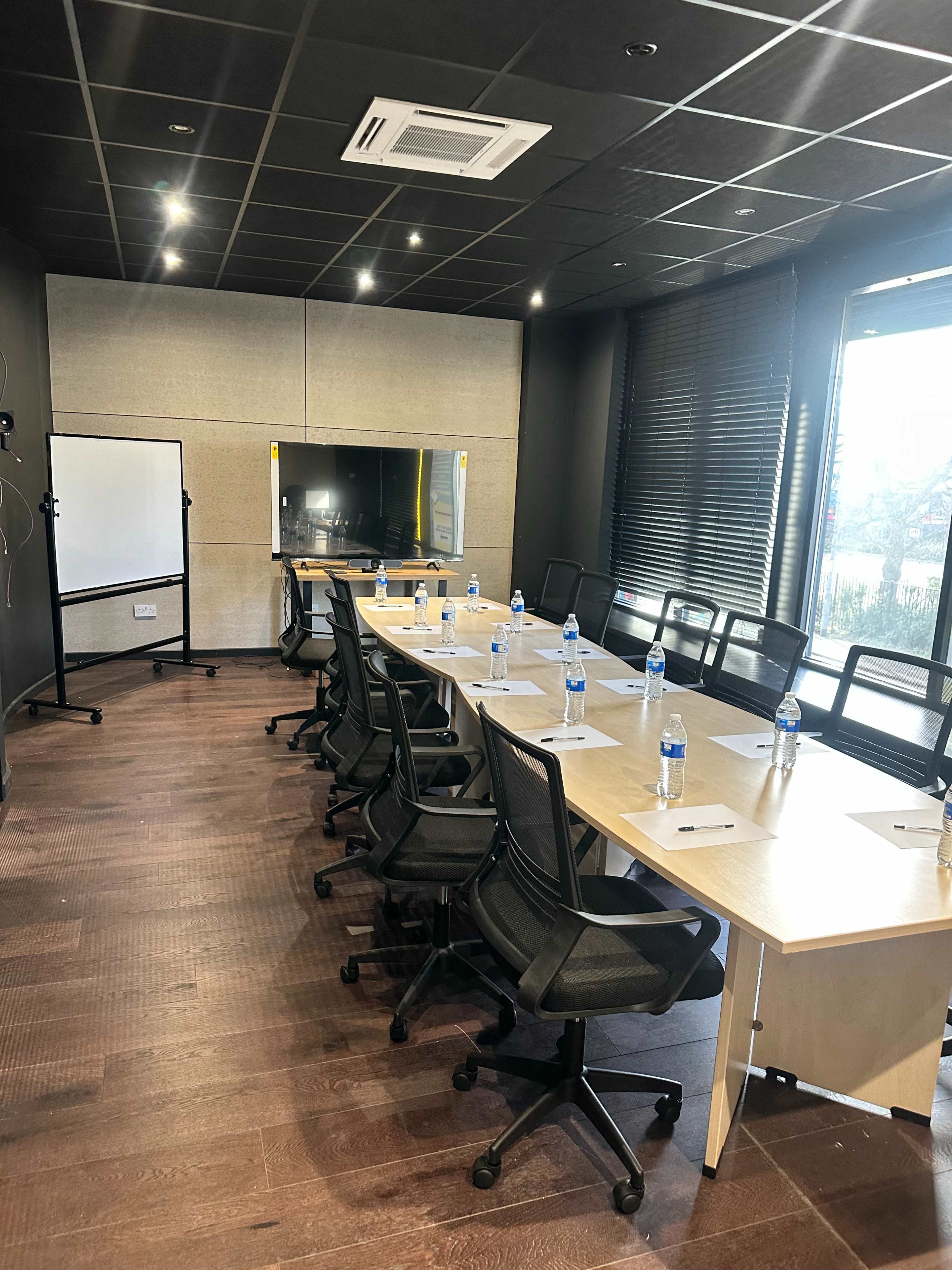 A modern conference room features a long table with chairs, equipped with water bottles and notepads, alongside a large screen and a whiteboard.