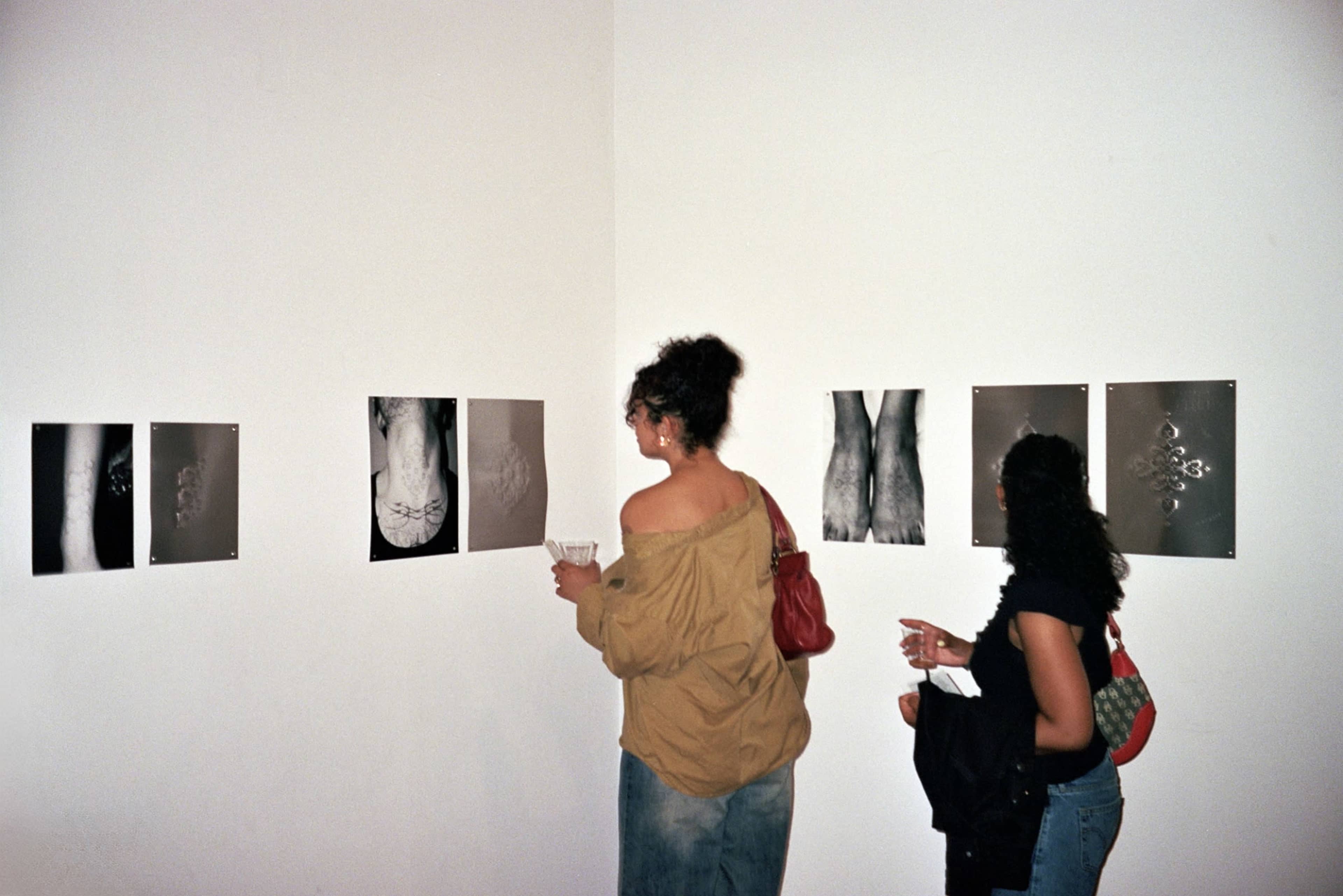 Two women observe black-and-white photographs displayed on a white wall in an art gallery.