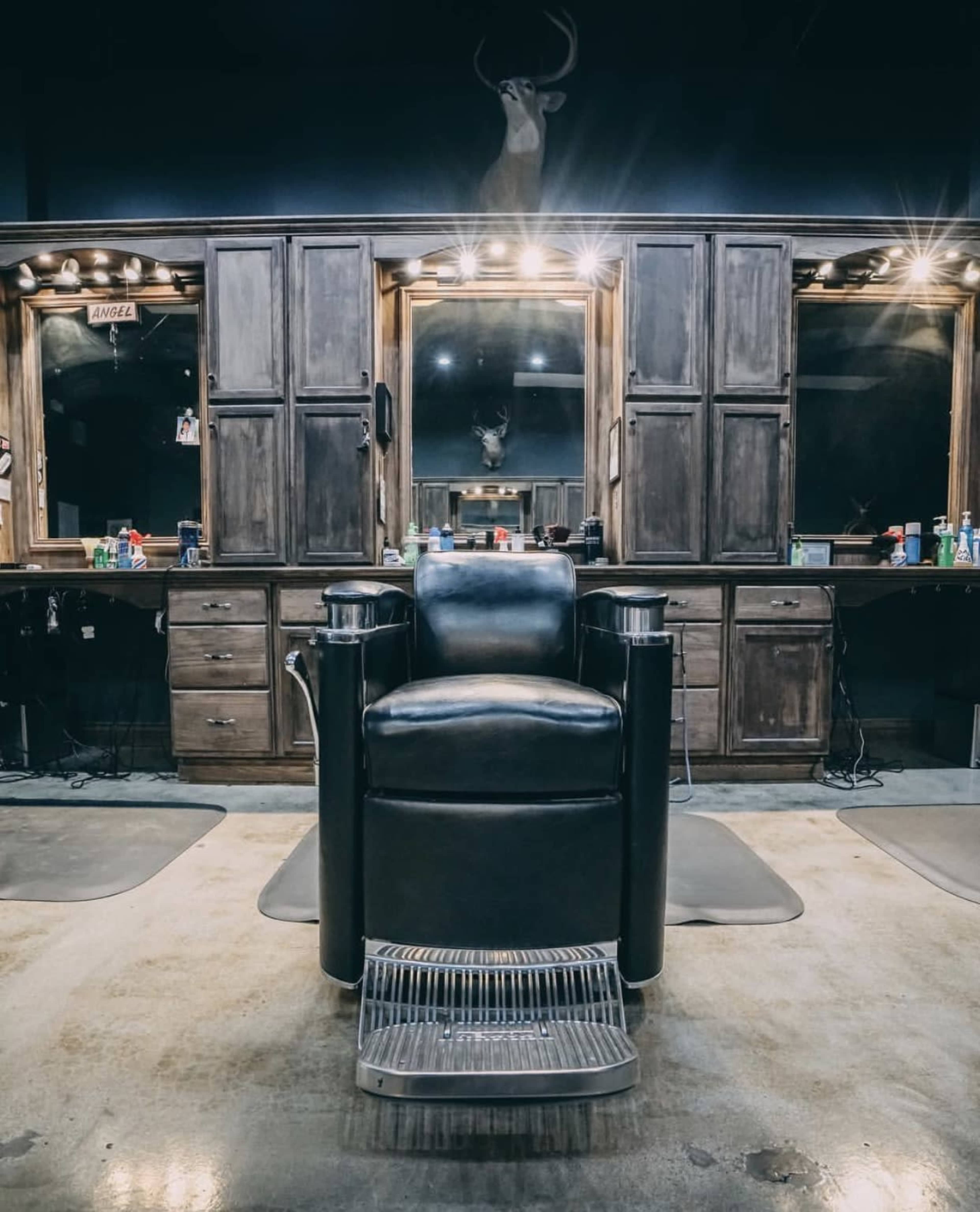 A black barber chair in front of a large mirror and wooden cabinetry in a dimly lit barbershop.