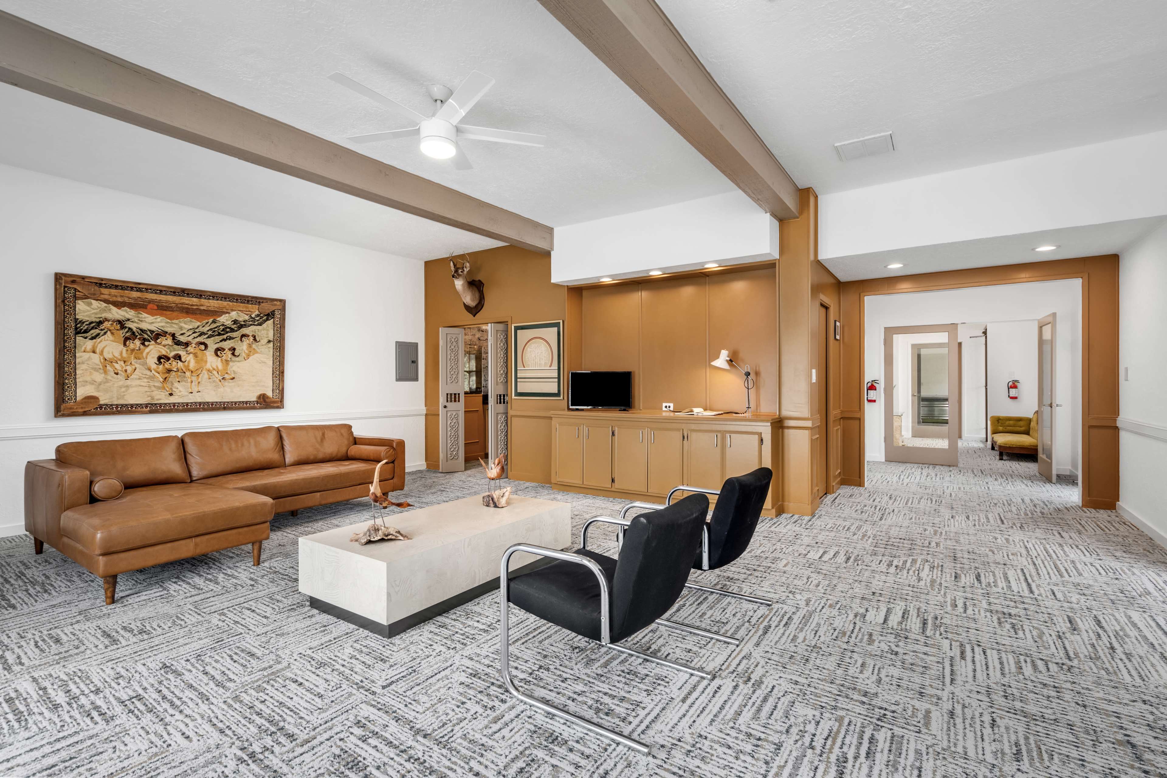 The image shows a modern living space featuring a brown leather sofa, black chairs, a white coffee table, and wooden beams along the ceiling.