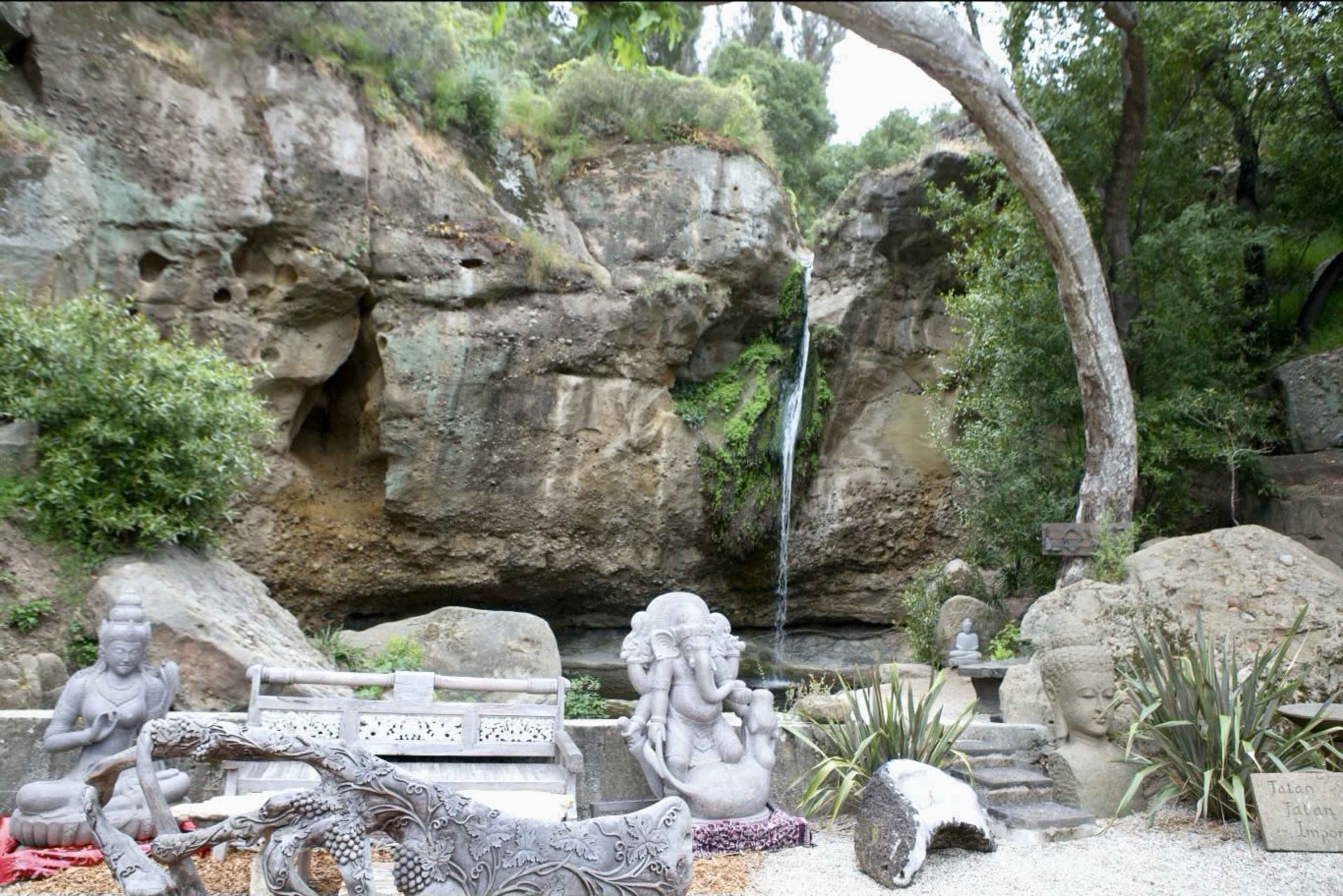 The image features a serene outdoor scene with stone sculptures, a small waterfall cascading from a rocky cliff, and lush greenery surrounding the area.