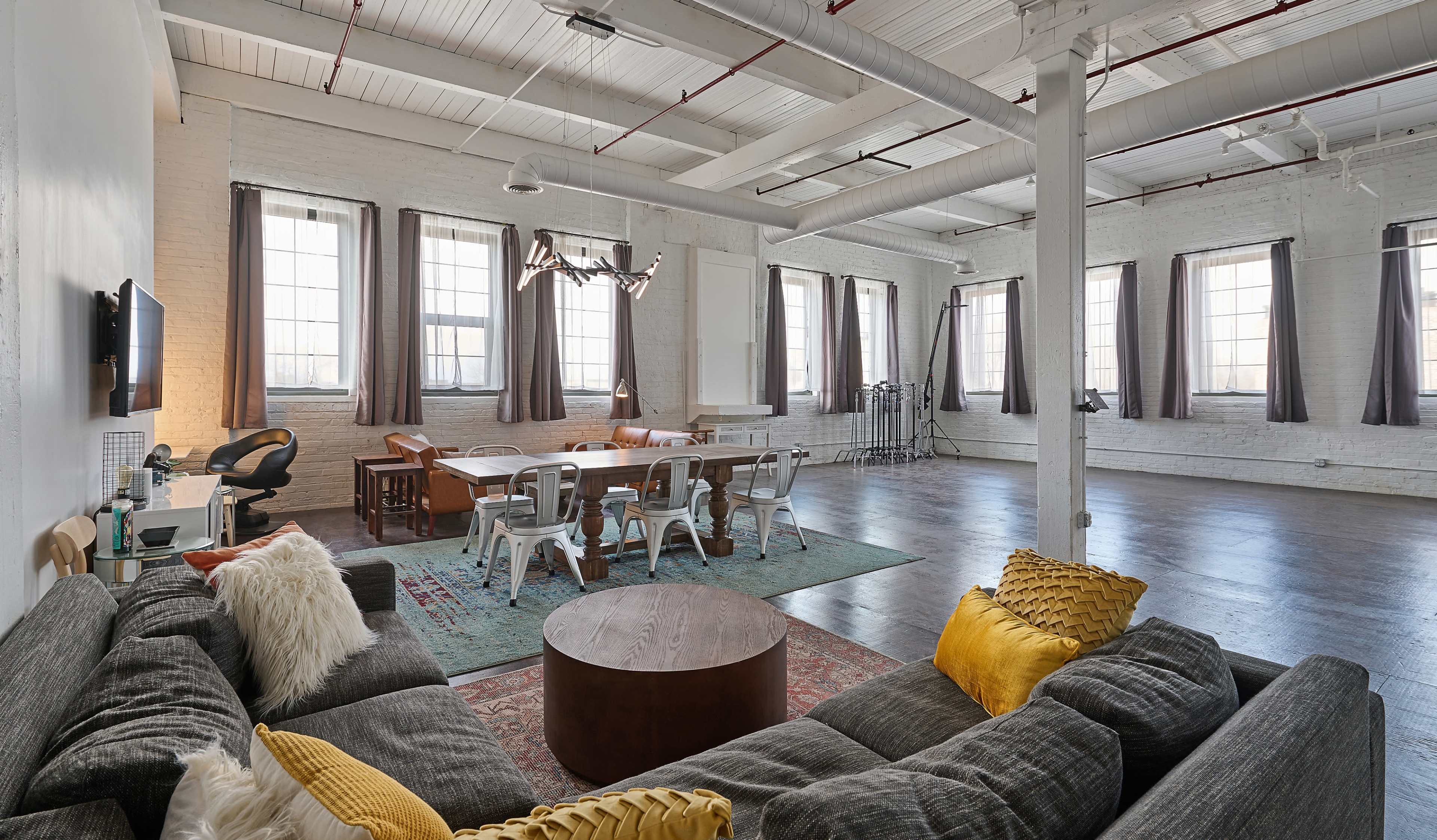The image shows a spacious, industrial-style loft with large windows, a seating area featuring a gray sofa and round coffee table, and a dining area with a wooden table surrounded by metal chairs.