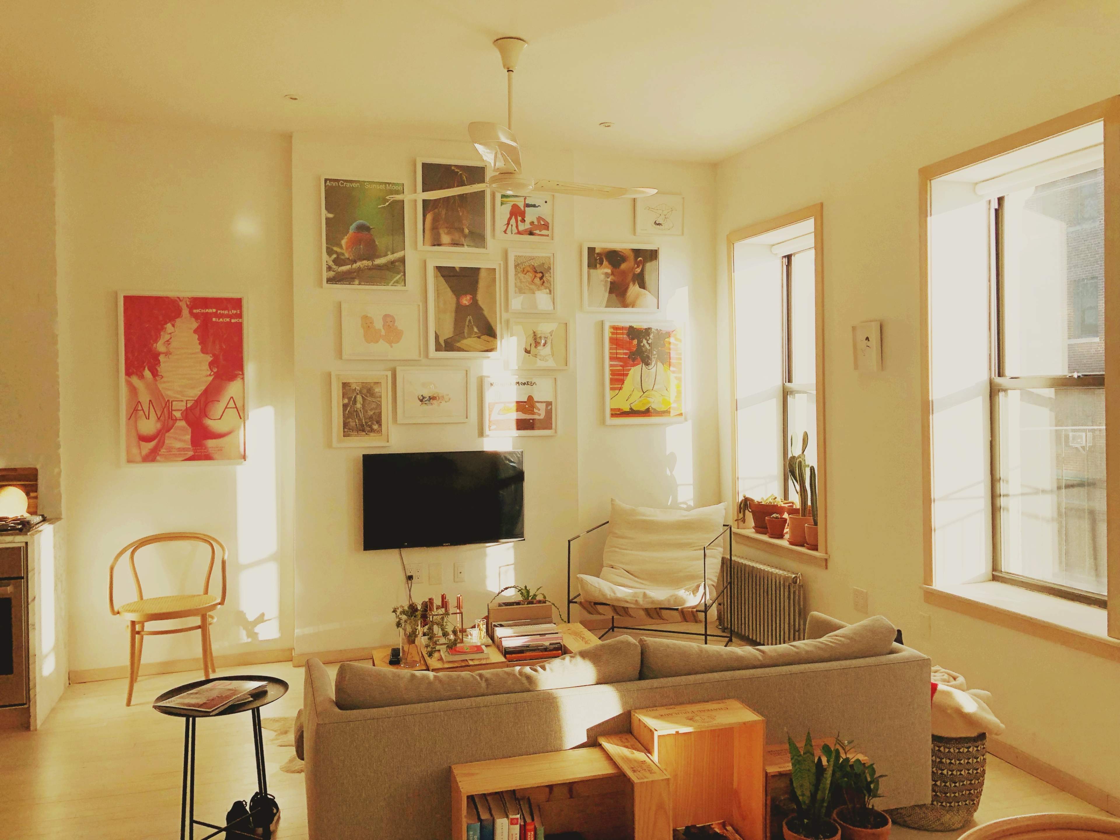 The image shows a brightly lit living room with a sofa, a wall adorned with various framed artworks, and large windows letting in natural light.