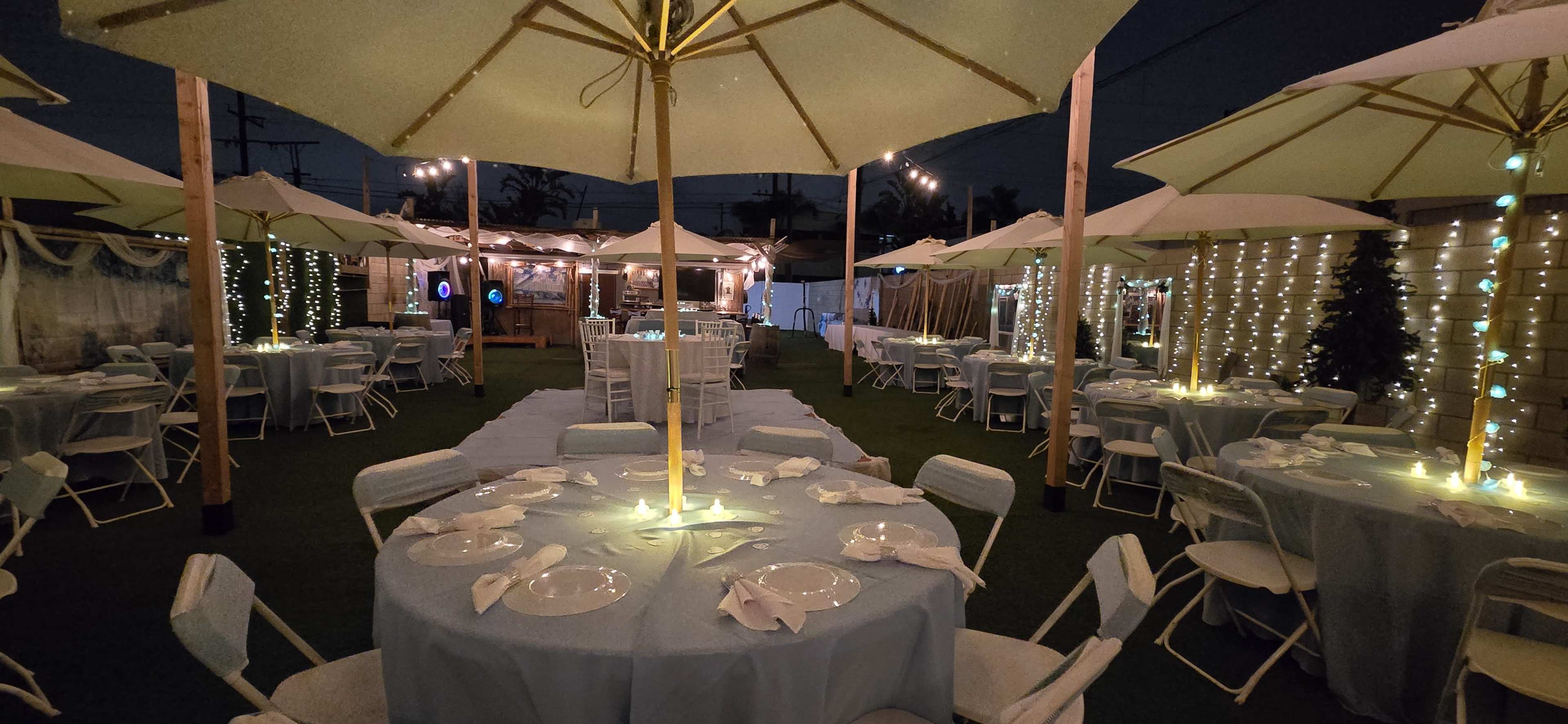The image shows a beautifully arranged outdoor event space with round tables covered in light blue tablecloths, each set with dinnerware and illuminated by soft lighting under white umbrellas.