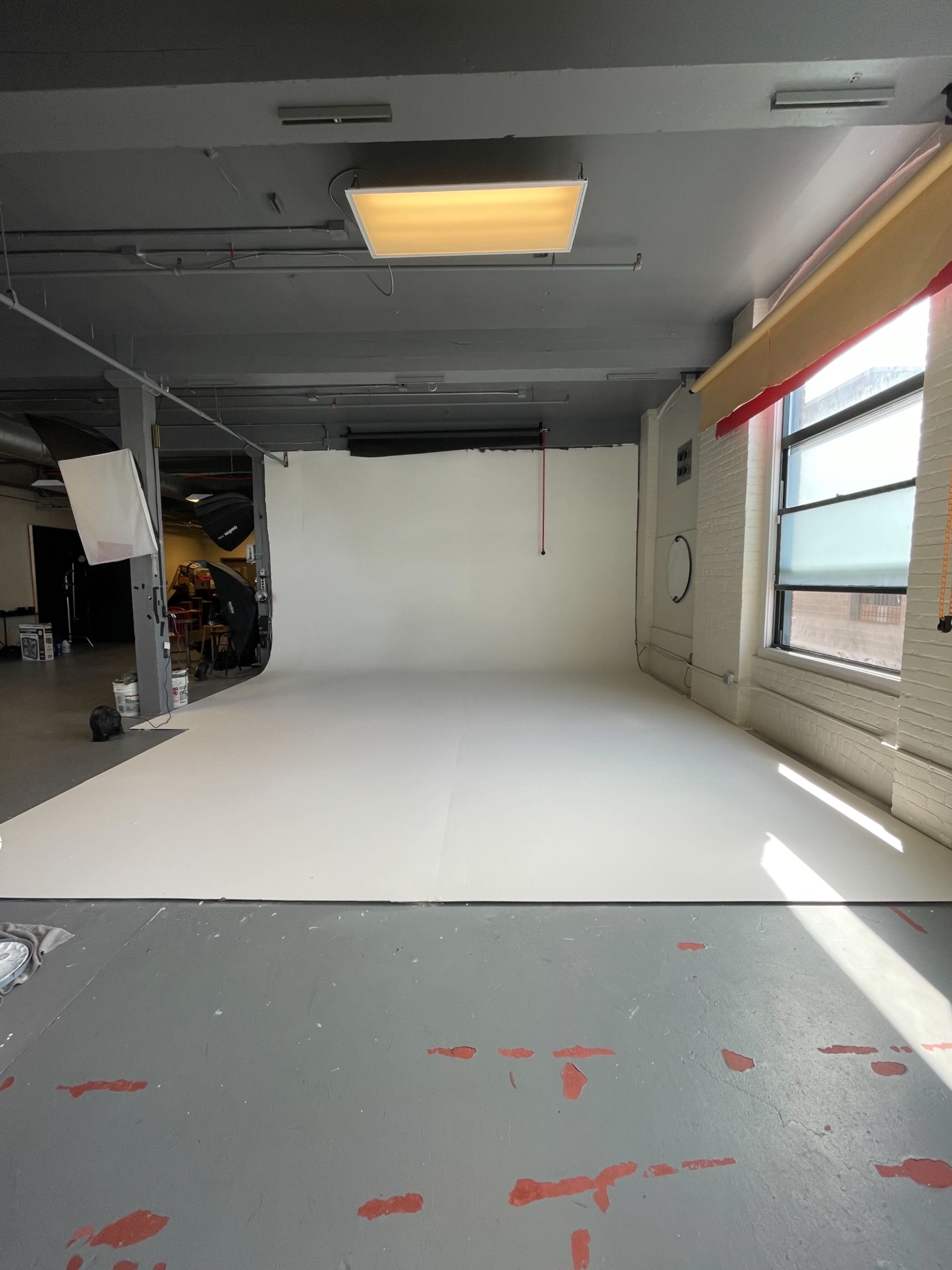 An empty photography studio with a white backdrop and gray flooring, illuminated by natural light from a large window.