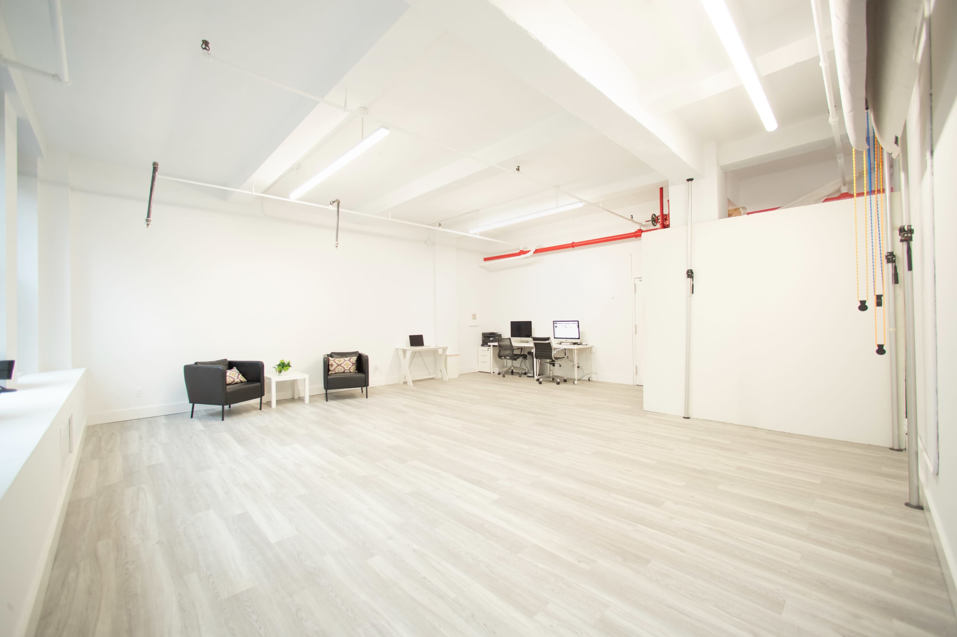 A bright, empty office space with light-colored wooden flooring, two black chairs, a desk with a computer, and white walls.