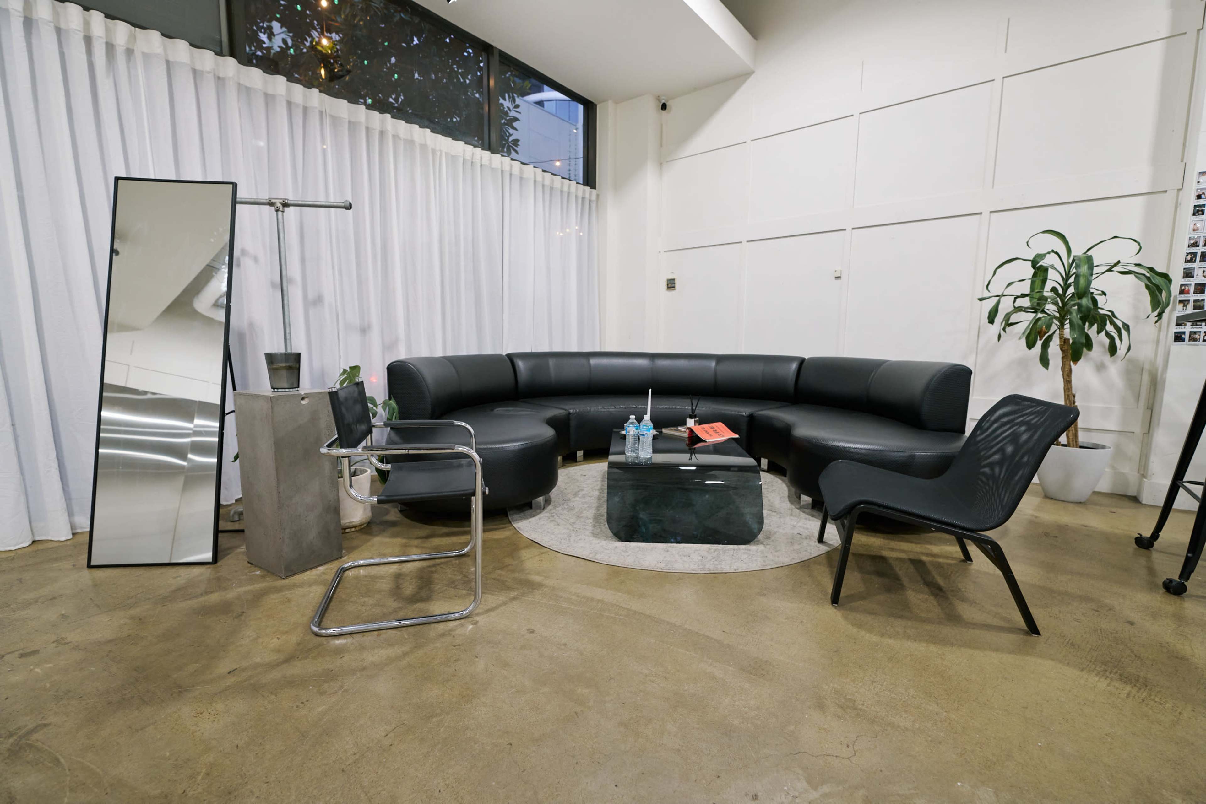 The image shows a modern seating area with a curved black sofa, a glass coffee table, a stylish chair, a floor mirror, and a plant, all set against a backdrop of white curtains and wall panels.