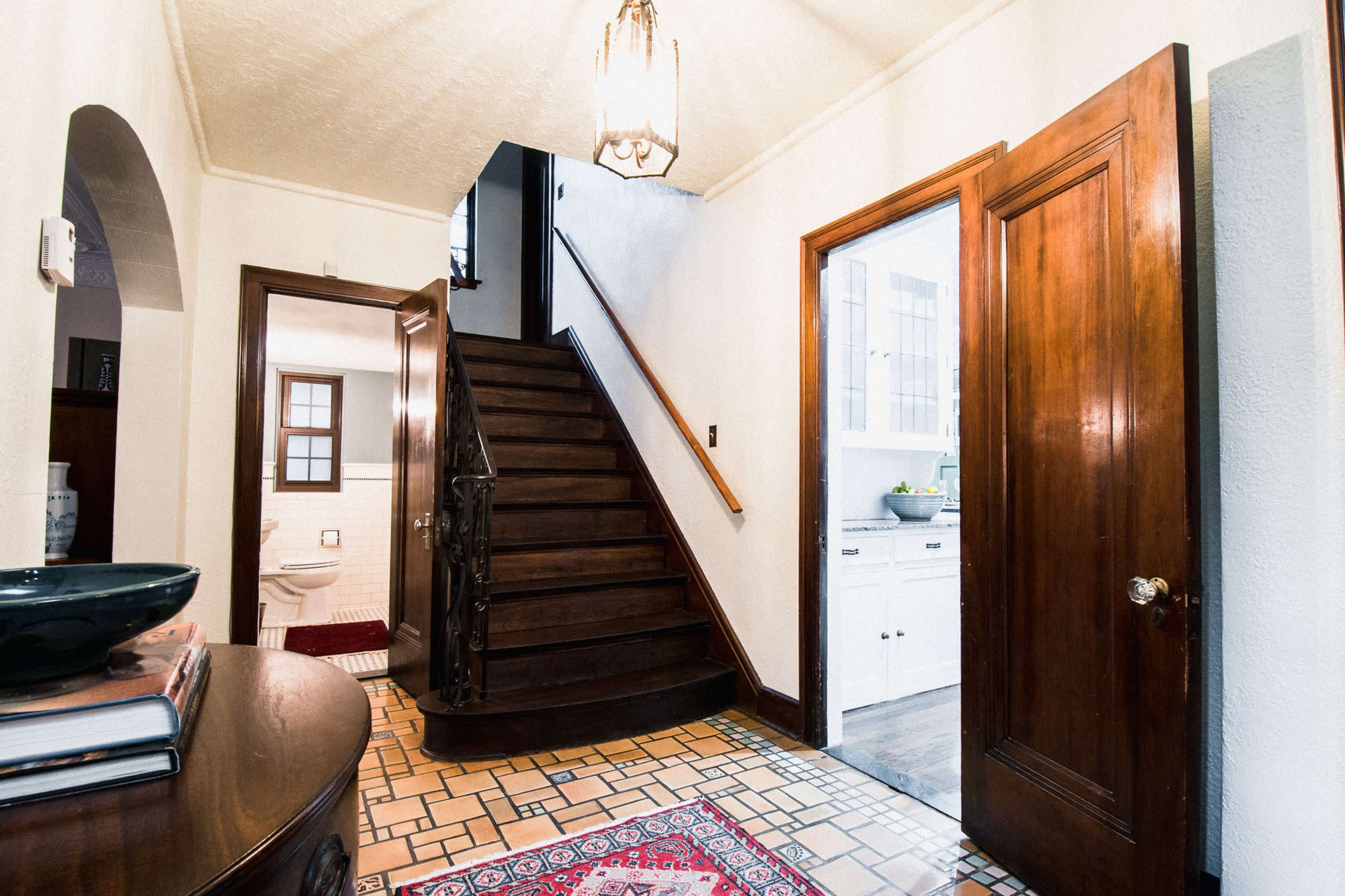 An entryway with a wooden staircase leading to an upper floor, a decorative rug on the tiled floor, and doorways leading to a bathroom and kitchen.