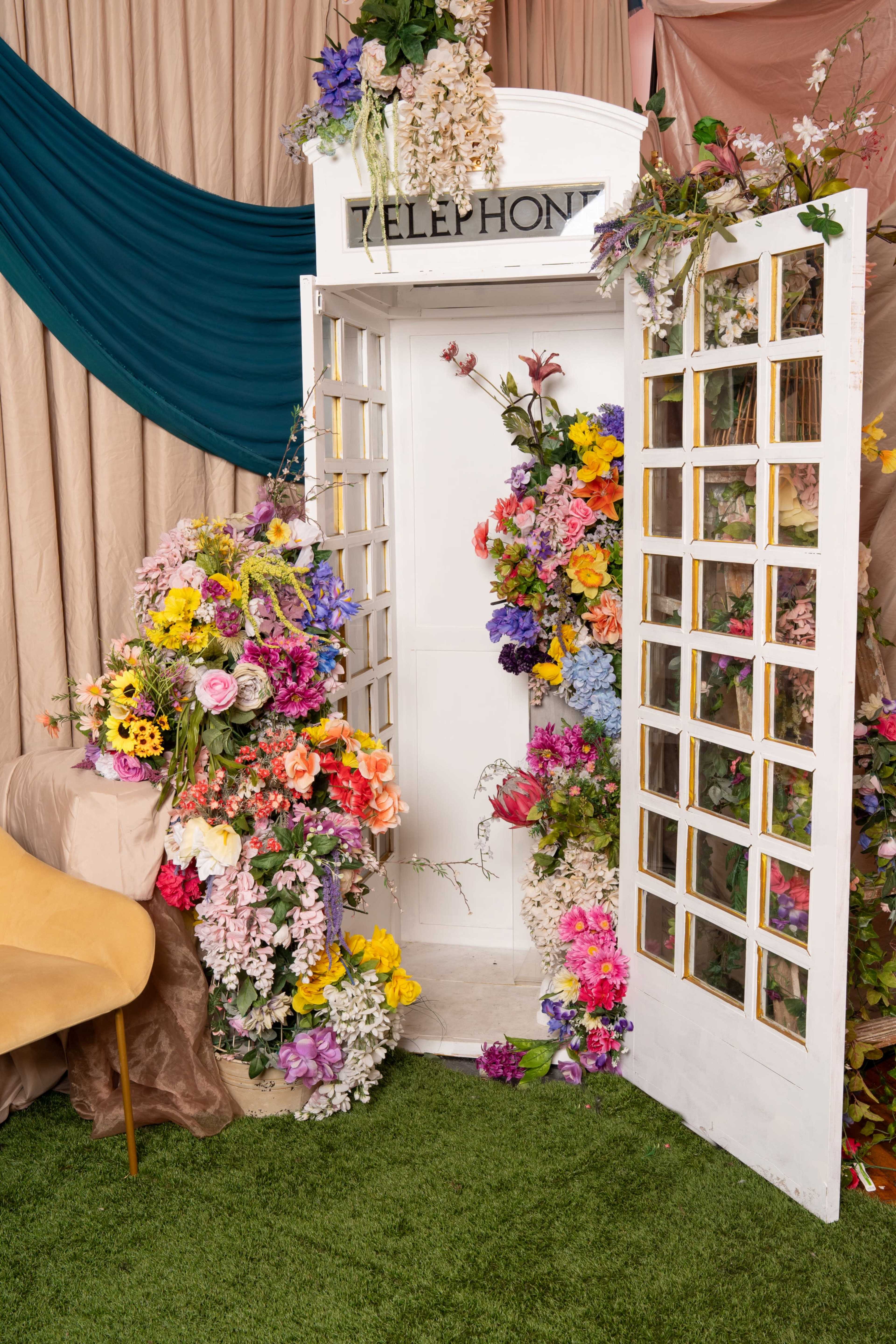 The image features a vintage telephone booth adorned with a variety of colorful artificial flowers, set against fabric drapes in a decorated indoor space.