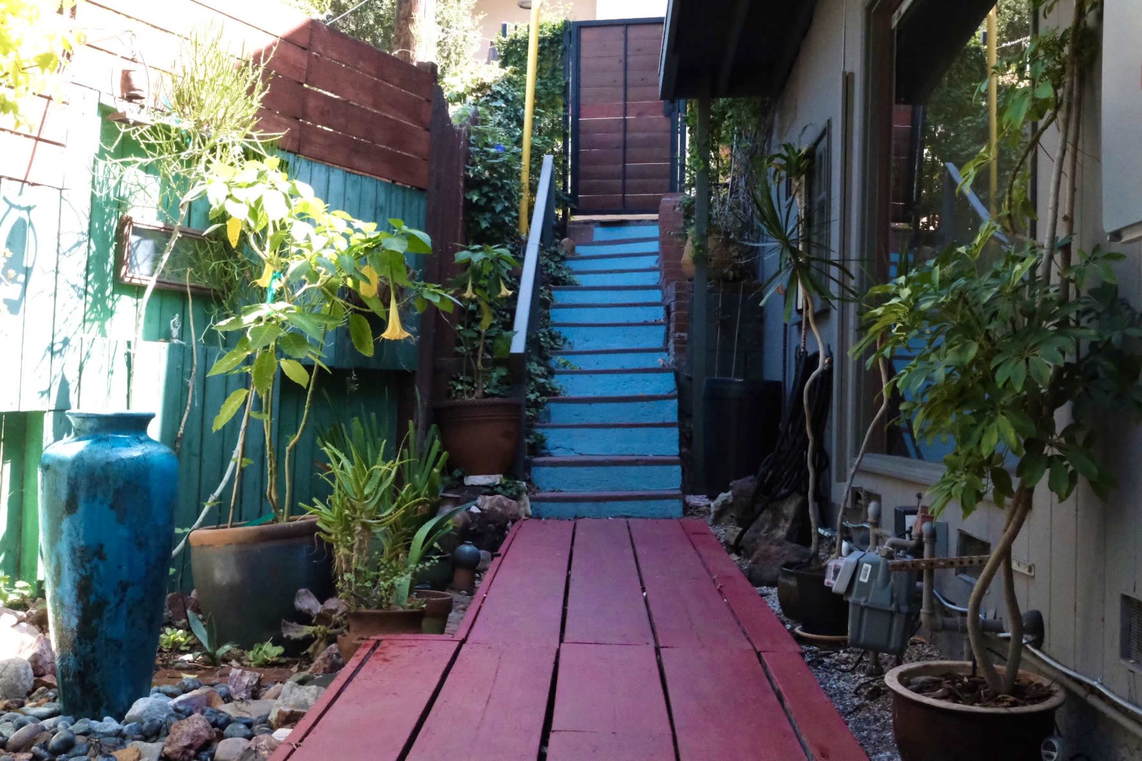 A narrow wooden walkway painted red leads to a set of blue stairs flanked by potted plants and surrounded by greenery.
