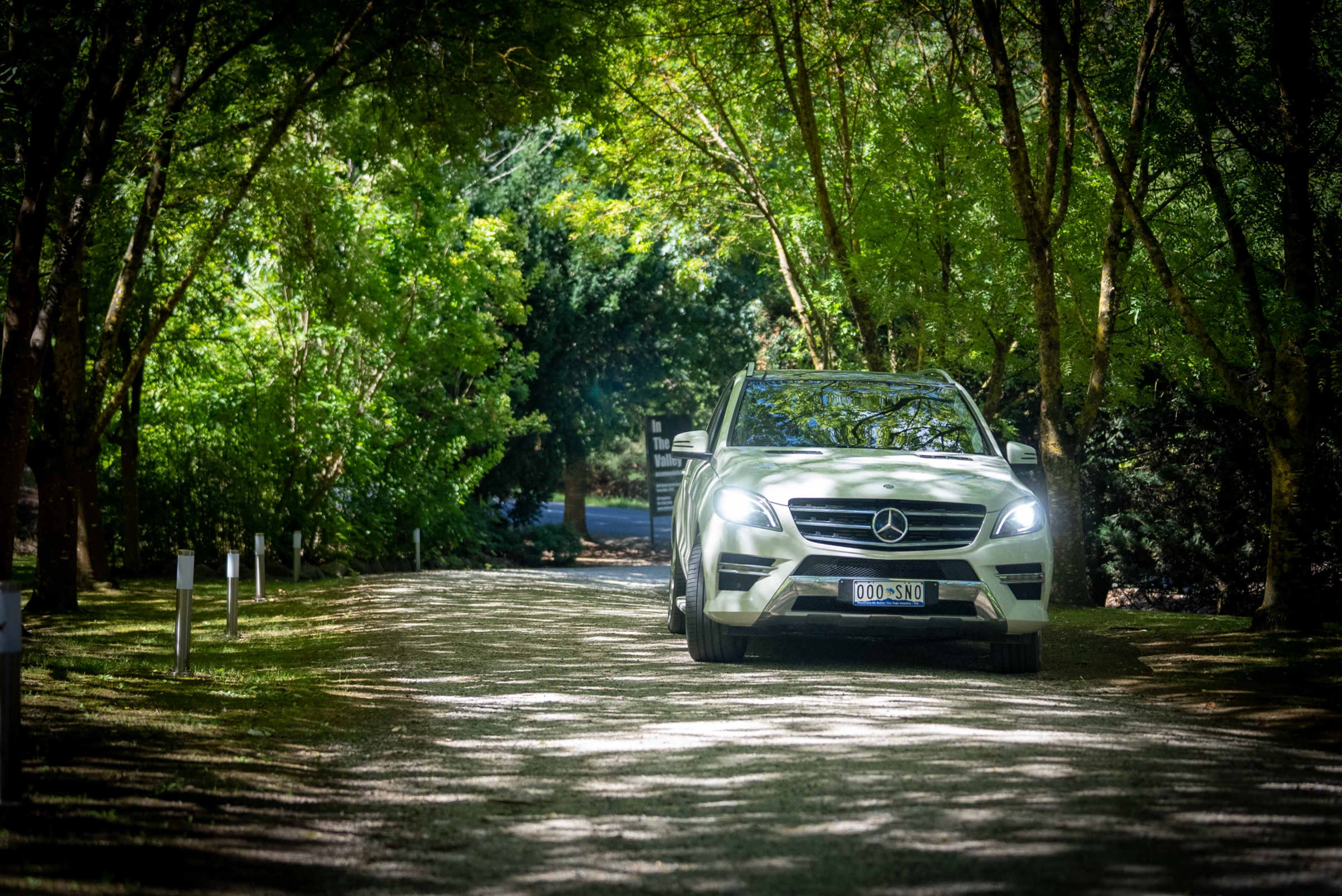 A white Mercedes-Benz SUV drives along a gravel path shaded by trees.