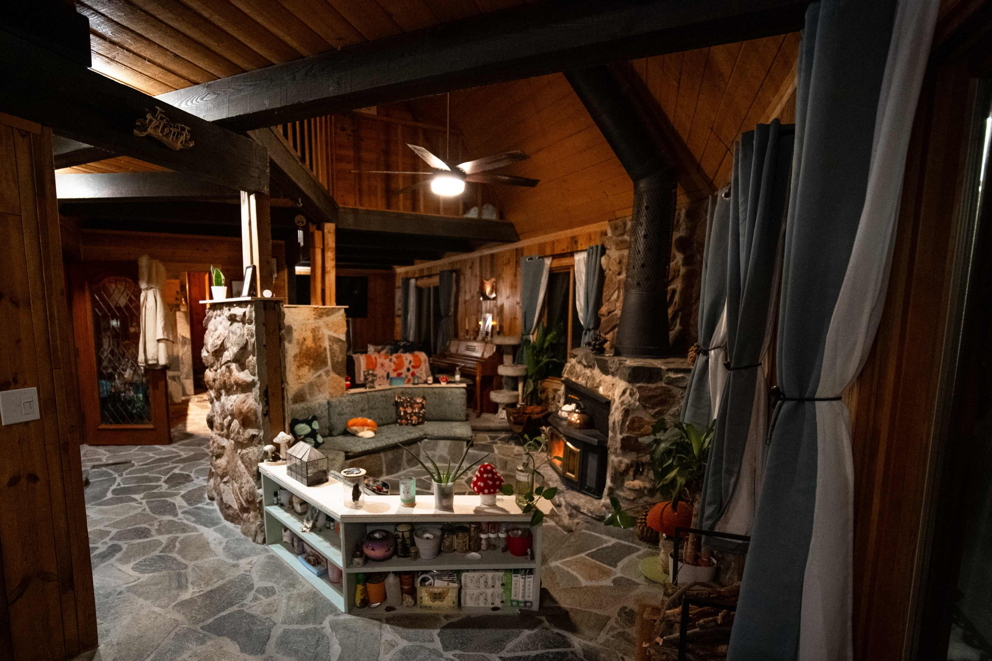 The image shows a cozy interior of a rustic living space featuring a stone-lined fireplace, a comfortable seating area, and wooden beams.