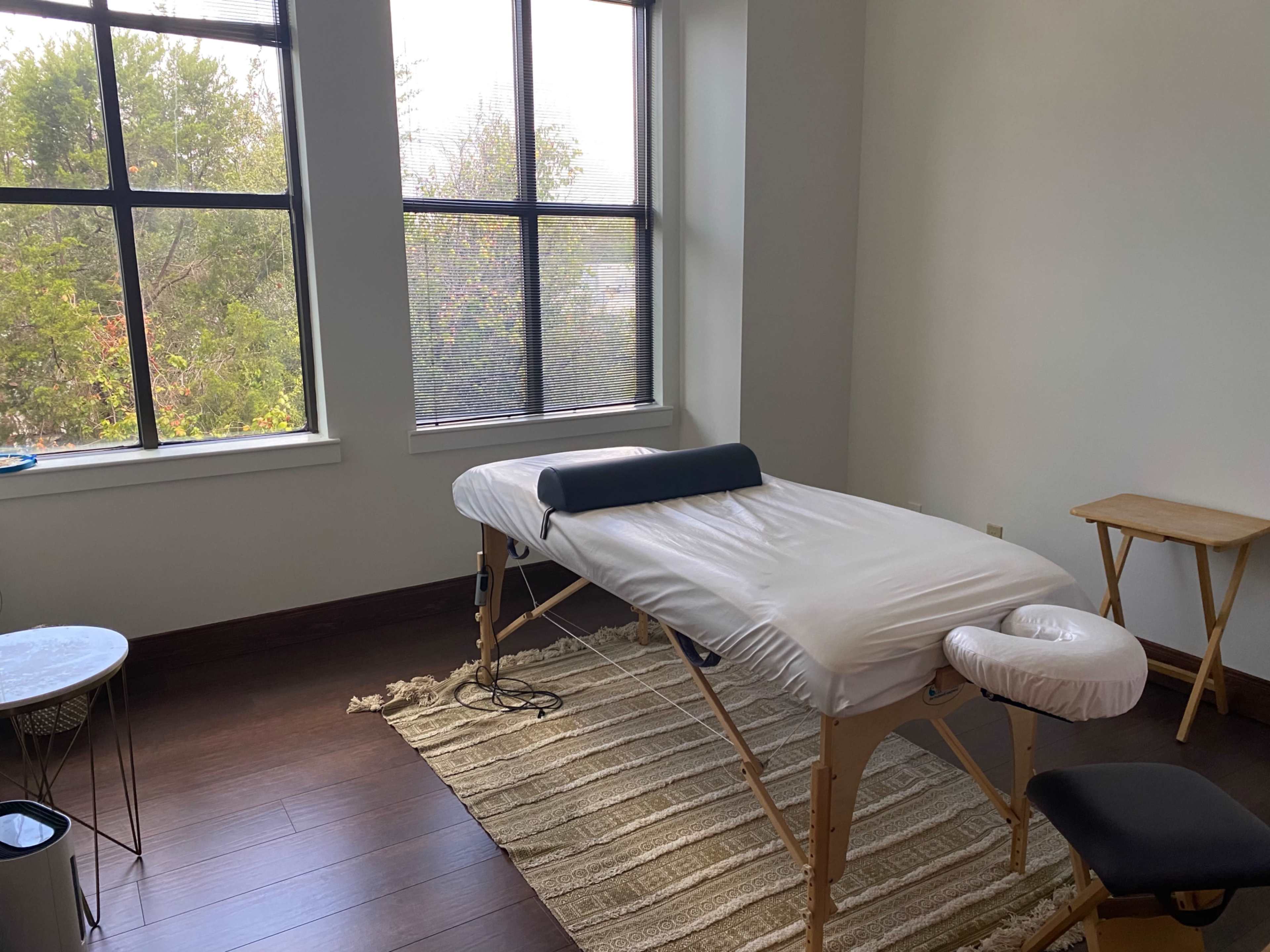 A massage therapy room features a massage table, a chair, and a small side table, all positioned under large windows with natural light.