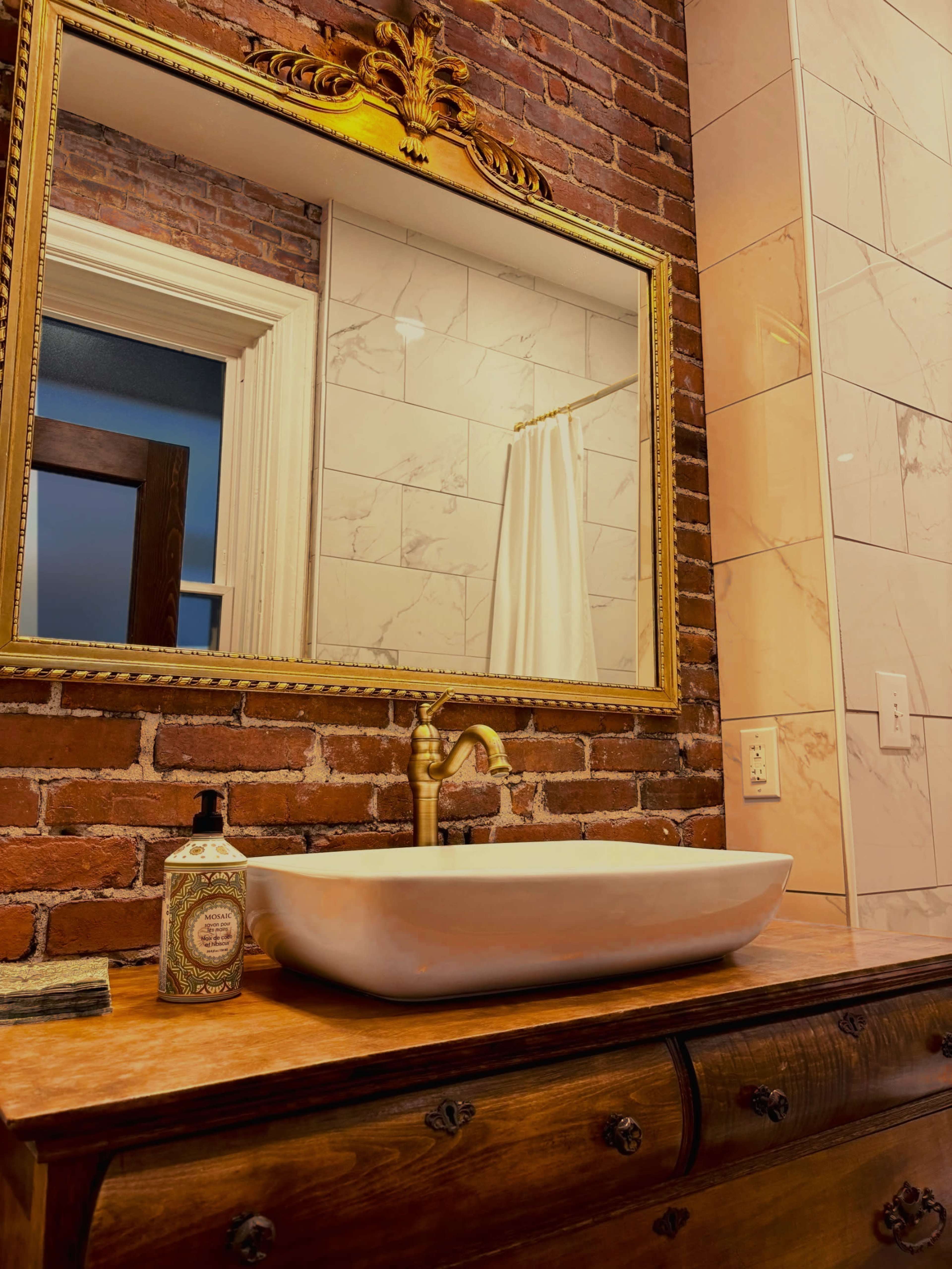 The image showcases a bathroom with a rustic brick wall, a large ornate mirror, and a white vessel sink atop a wooden vanity.