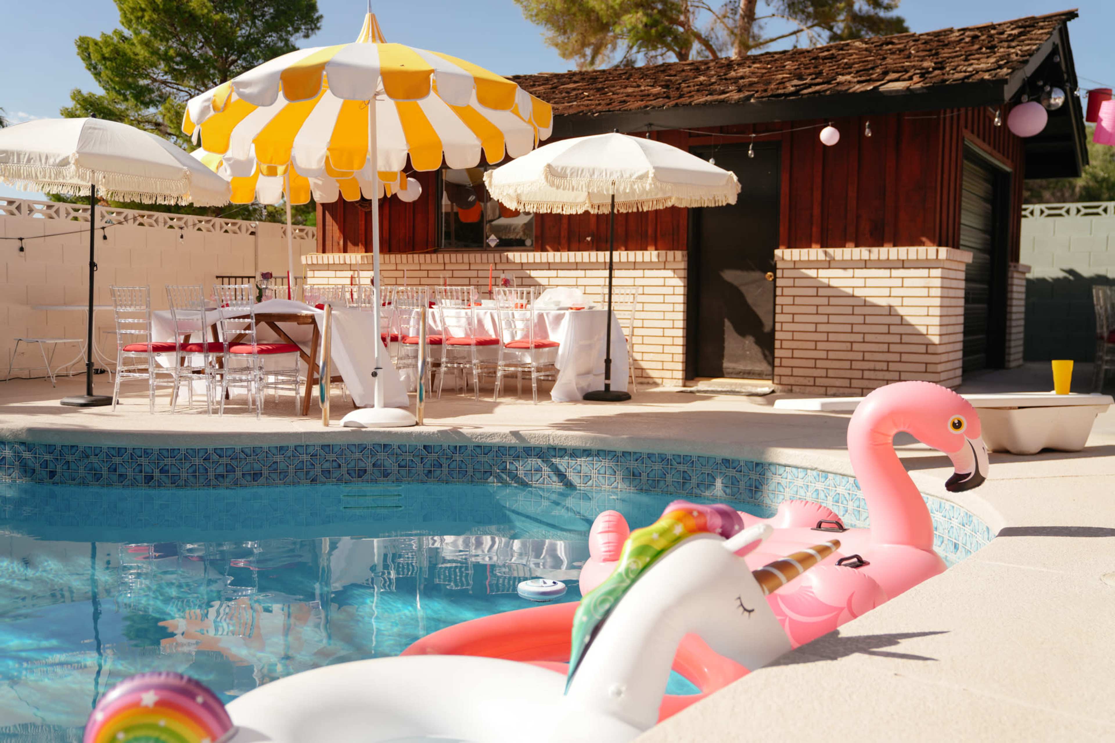 The image shows a swimming pool with inflatable flamingo and unicorn toys, surrounded by a patio featuring a striped umbrella and a decorated dining area.