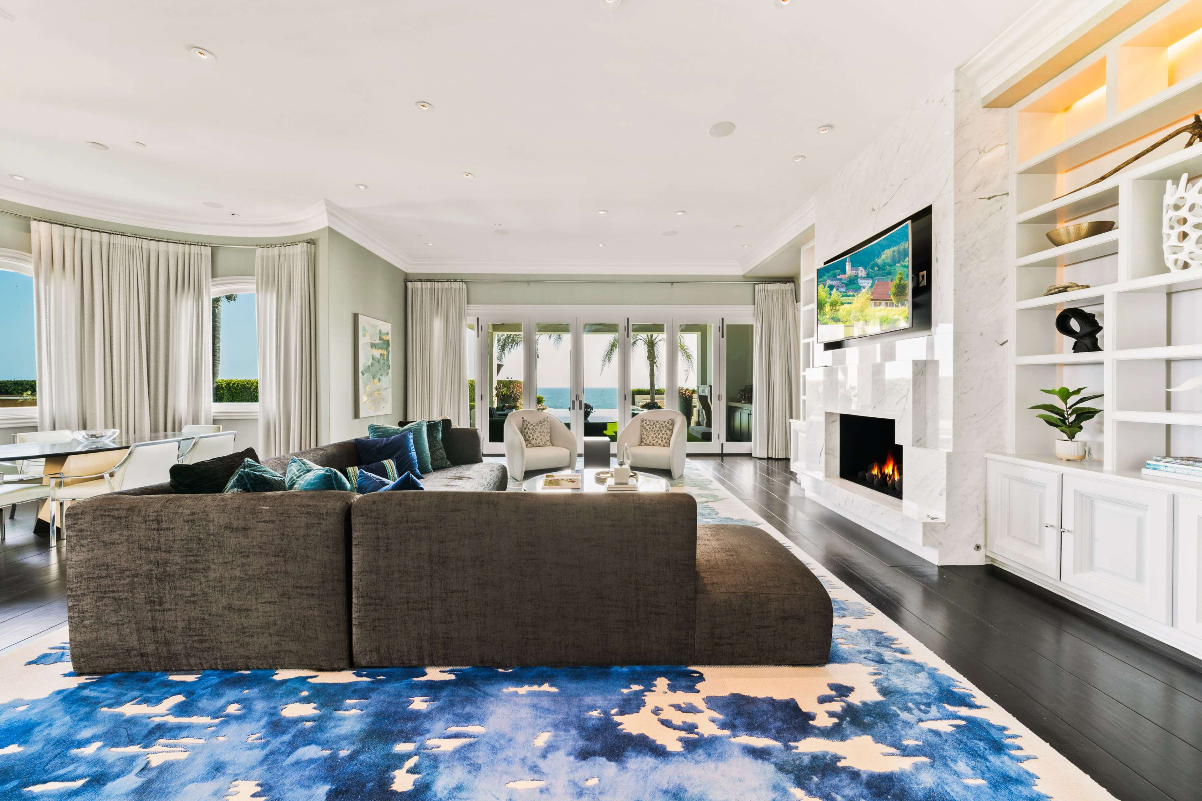 The living room features a large sectional sofa, a modern fireplace, a wall-mounted television, and large windows that offer a view of the outdoors.