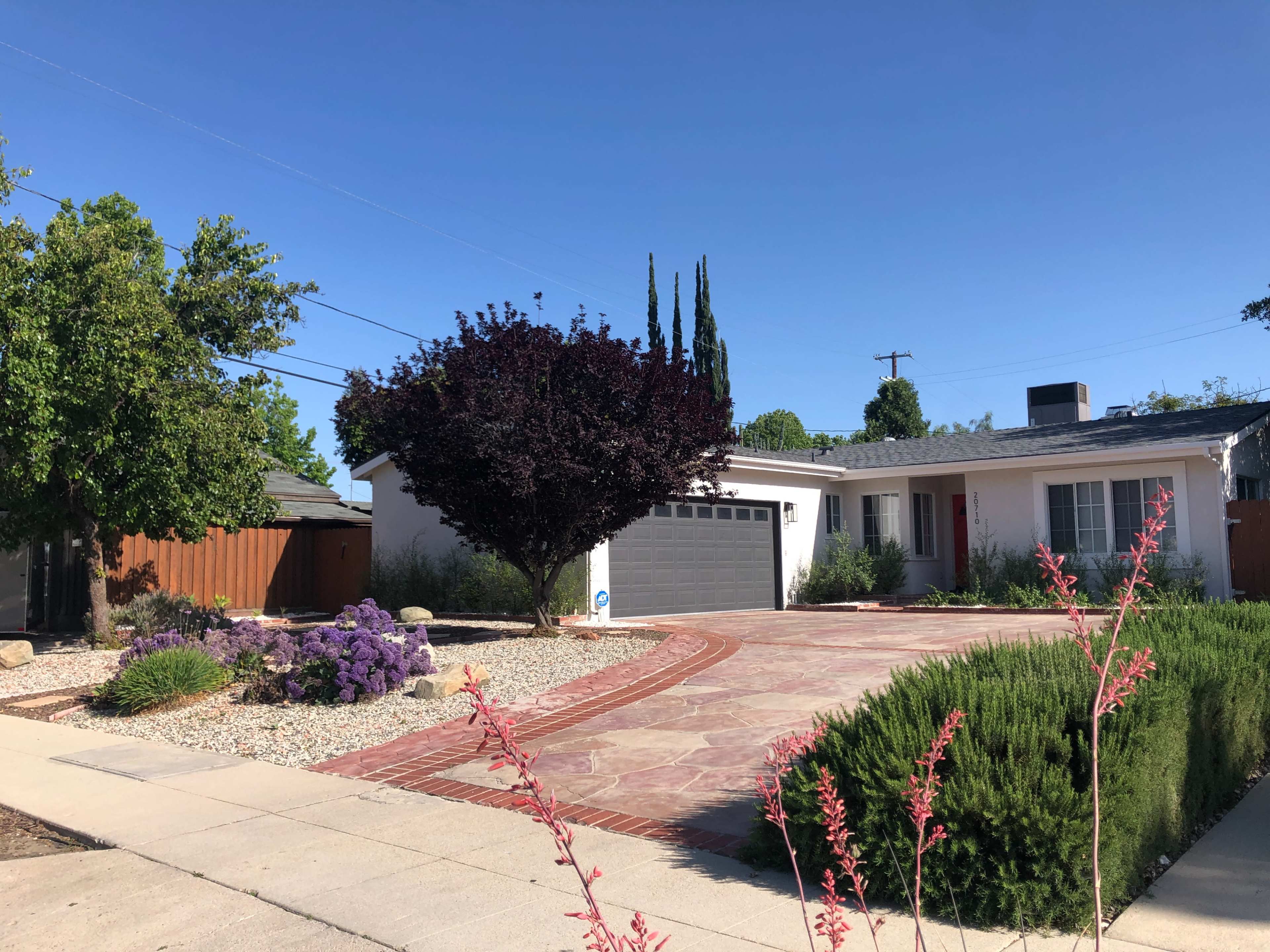 The image shows a residential house with a driveway made of patterned stone, surrounded by landscaped plants and a large purple tree in the front yard.