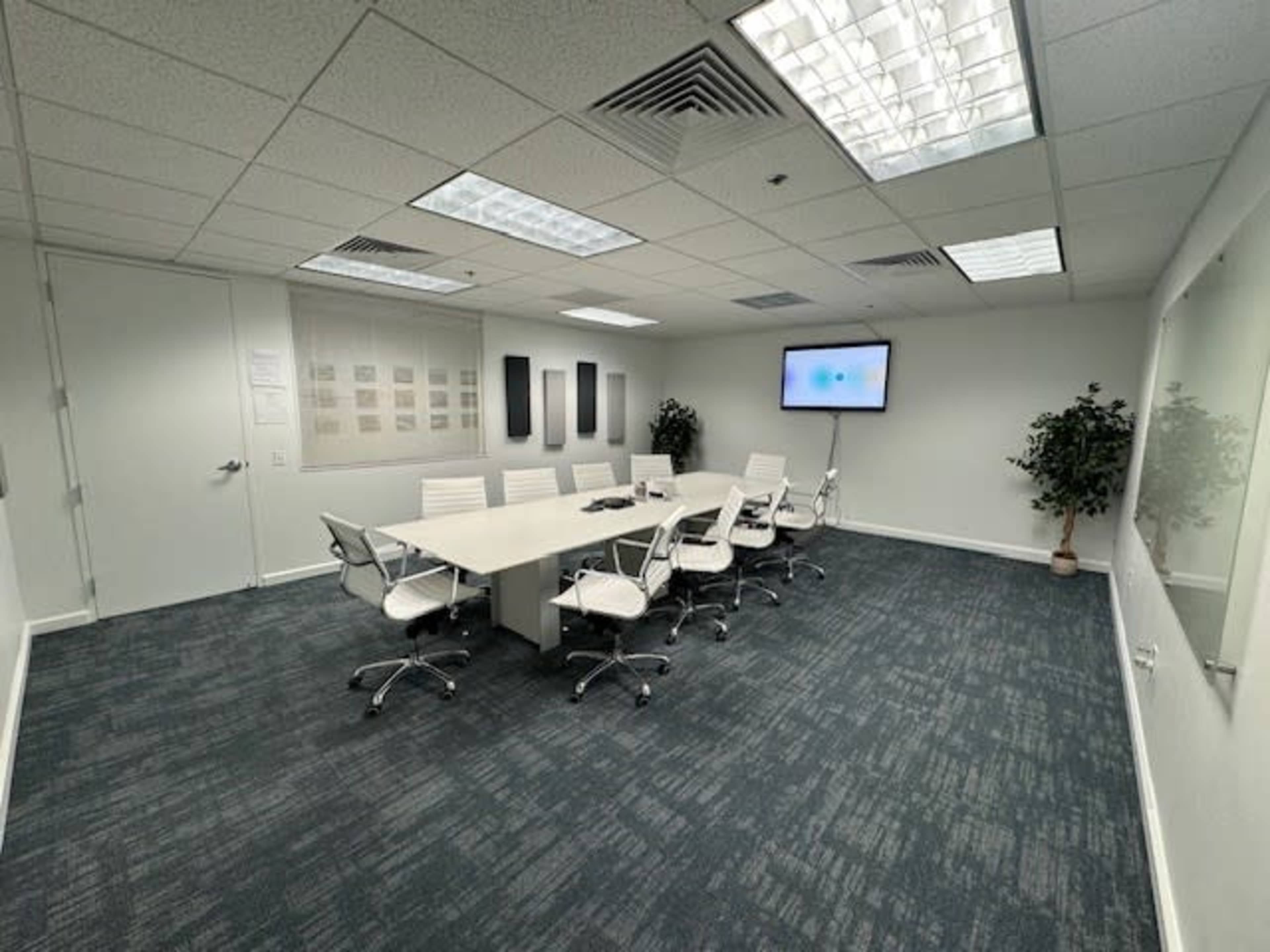 A modern conference room features a long table surrounded by white chairs, a large screen on one wall, and two potted plants in the corners.