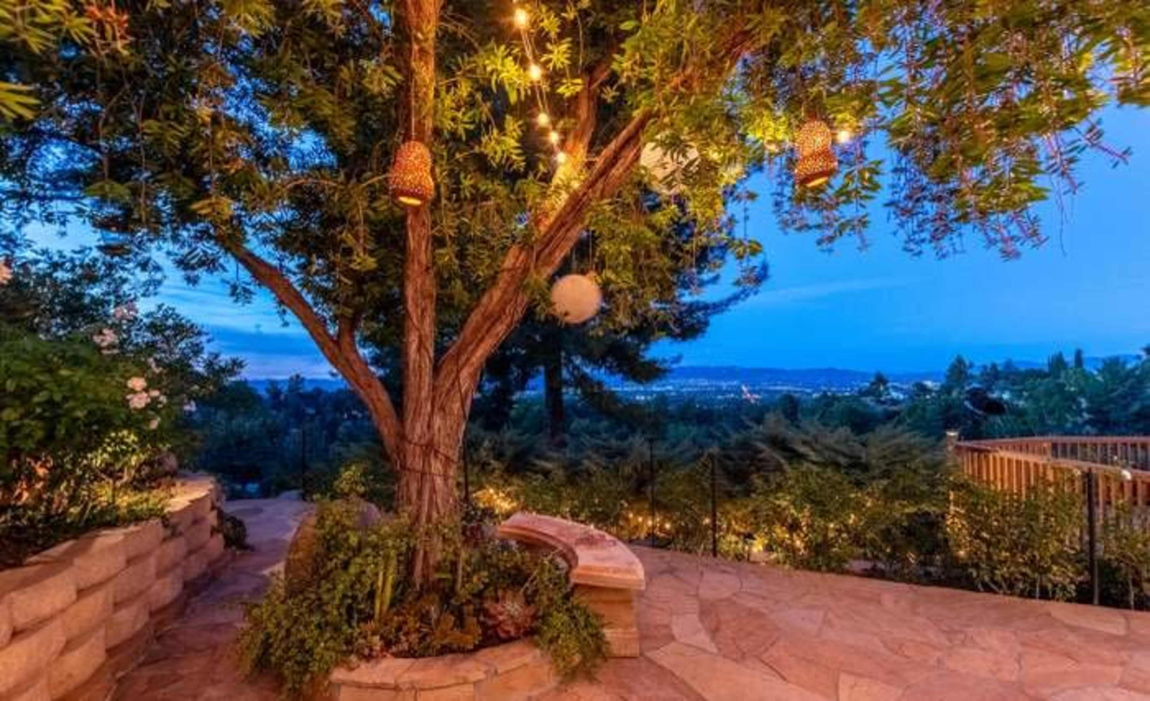 A tree adorned with decorative lights stands in a landscaped outdoor area, overlooking a distant view at dusk.
