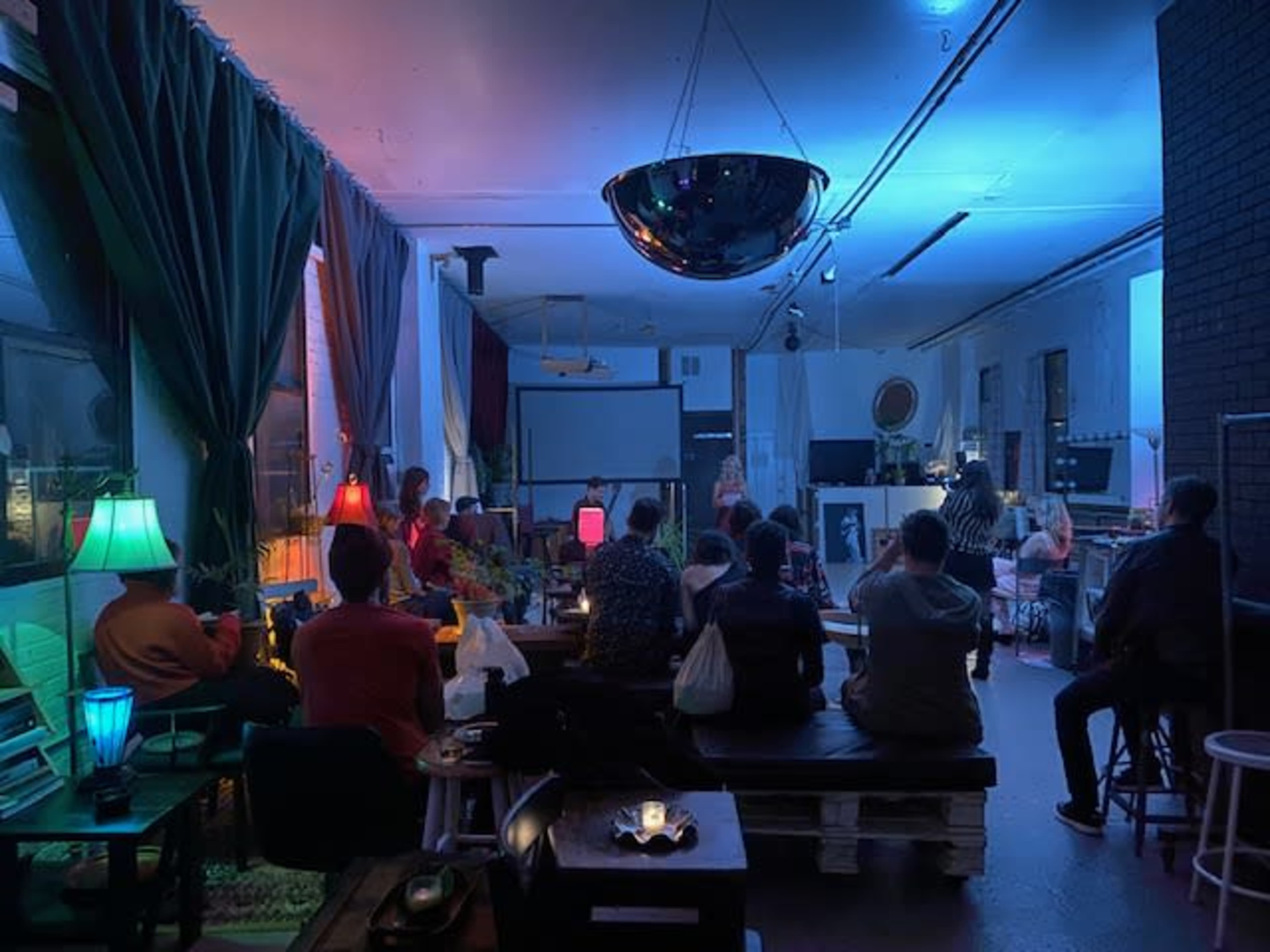 A group of people sit in a dimly lit room with colorful lamps during a gathering or presentation.