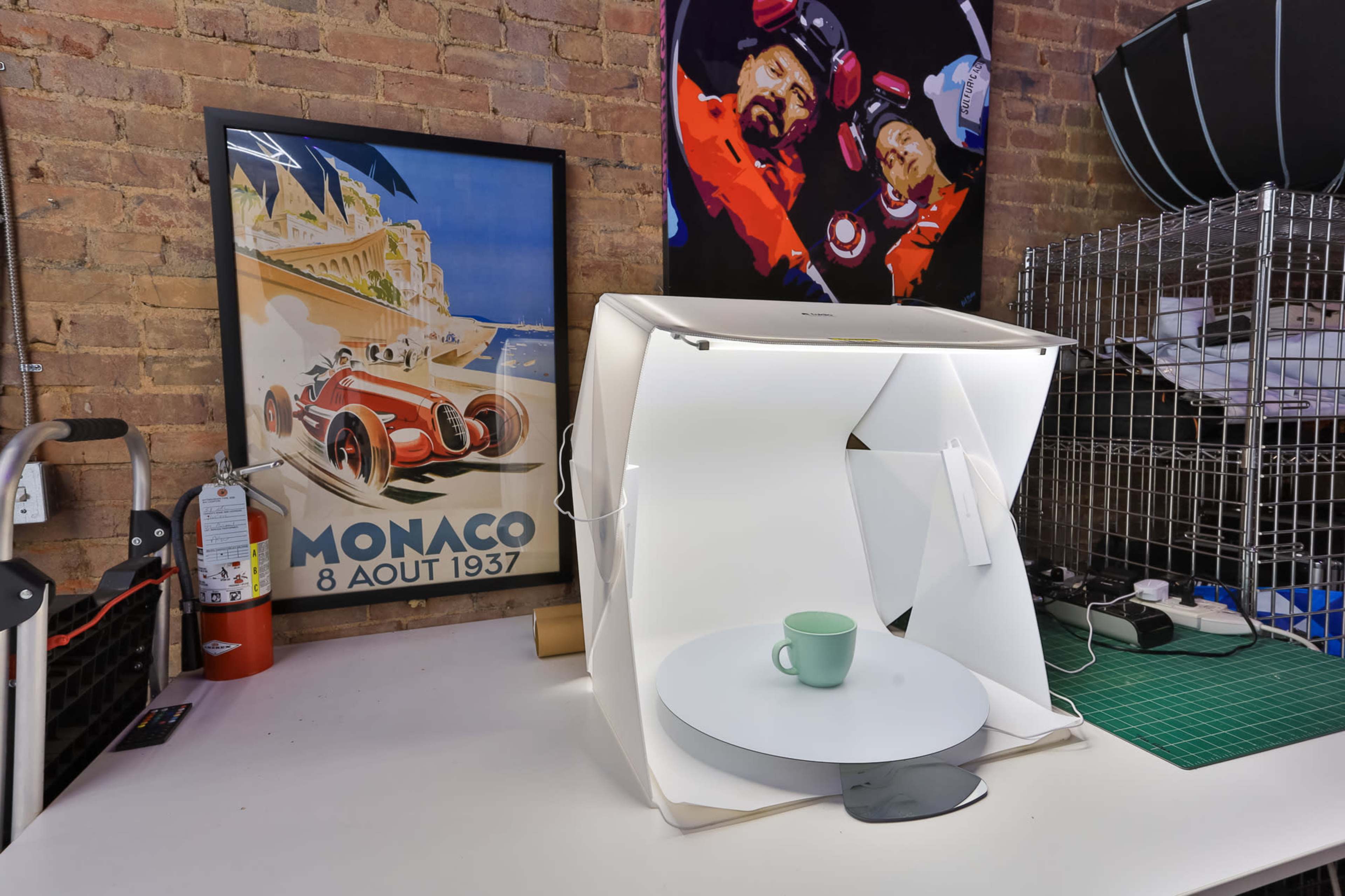A white photo studio tent is set up on a table beside a framed vintage poster of a car race in Monaco.