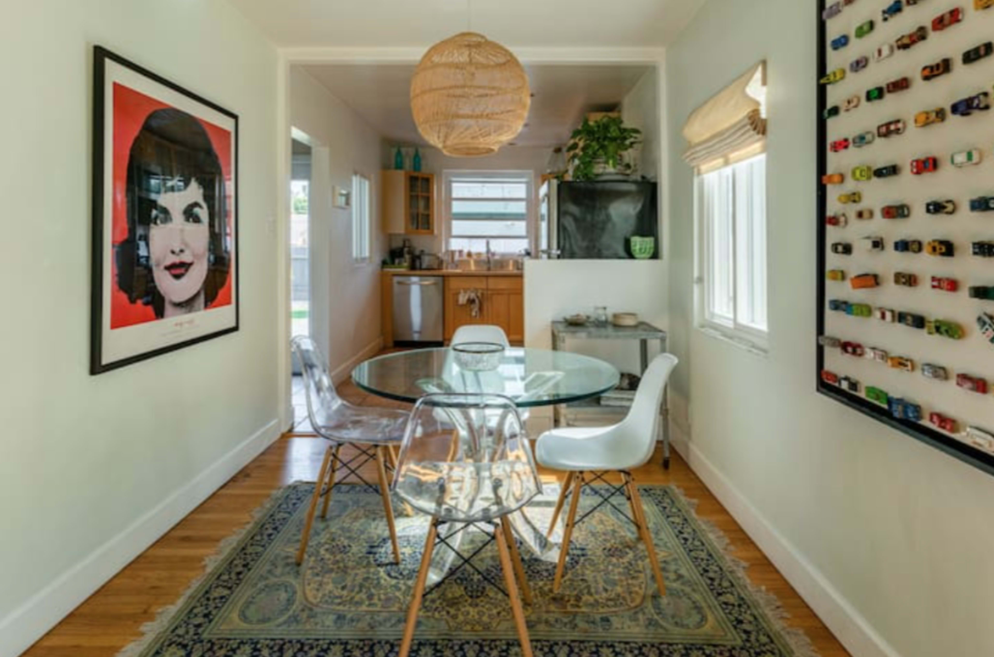 A glass dining table with four chairs is positioned in a hallway leading to a kitchen, featuring a large framed artwork and a wall display of toy cars.