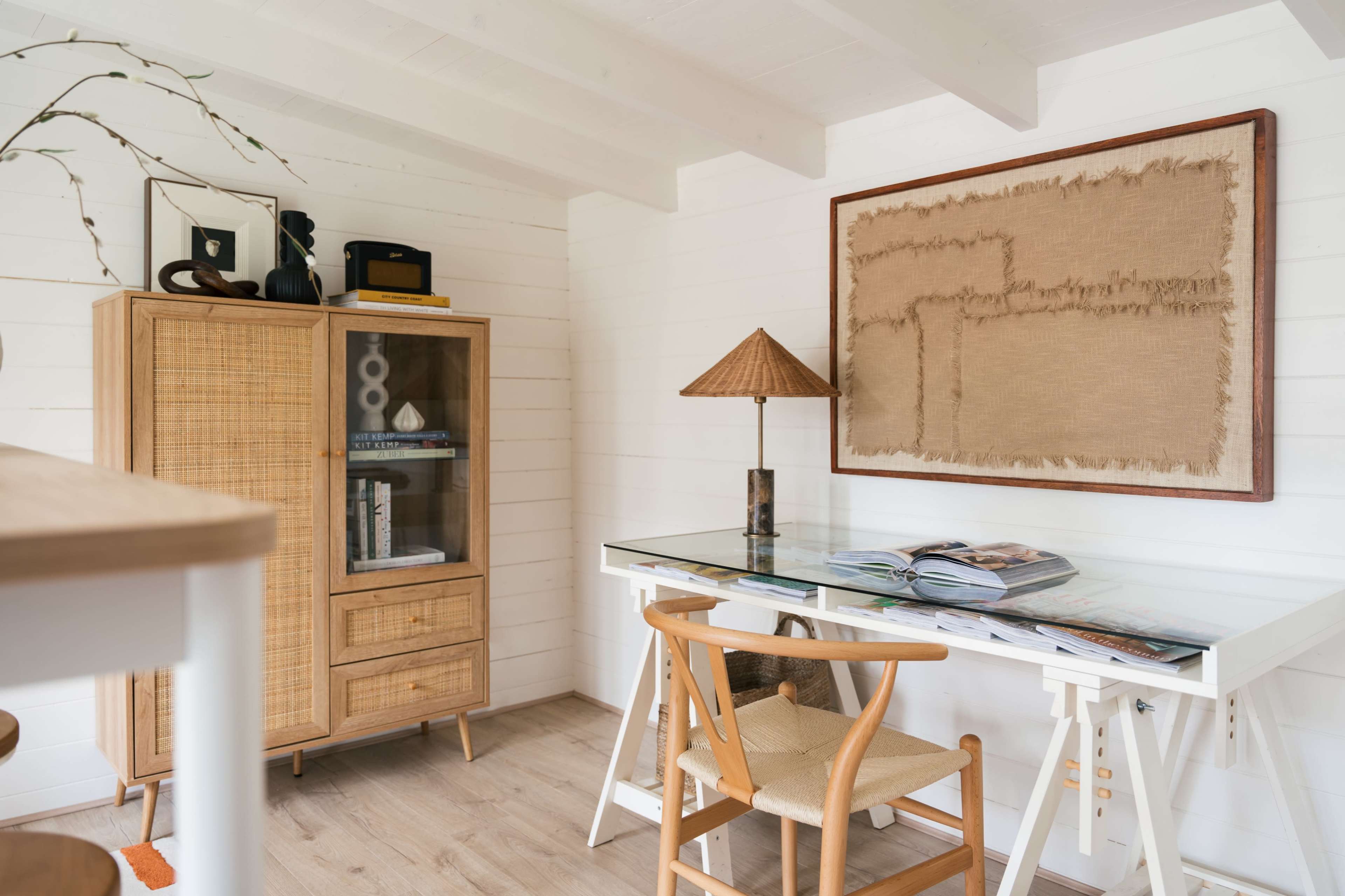 A wooden desk with a glass top, accompanied by a chair, is positioned next to a wicker cabinet in a well-lit room with white walls and wooden beams.