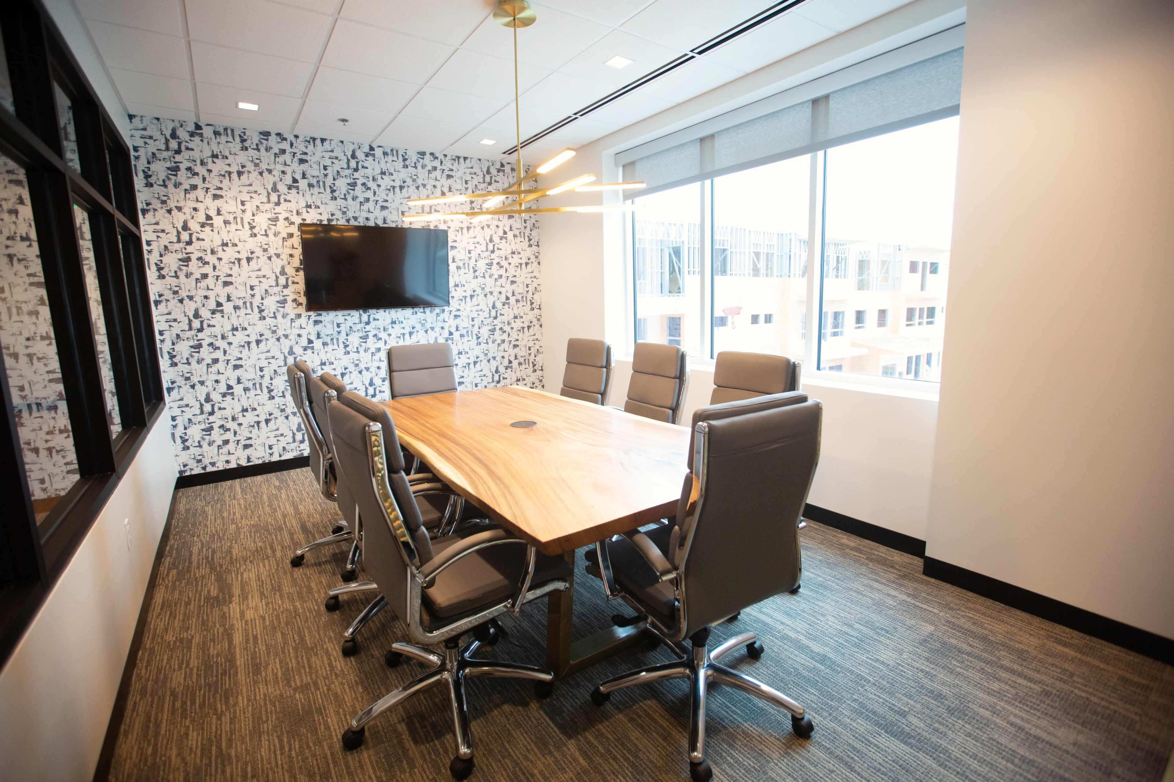 A conference room features a large wooden table surrounded by eight gray office chairs, with a television mounted on the wall and windows providing natural light.