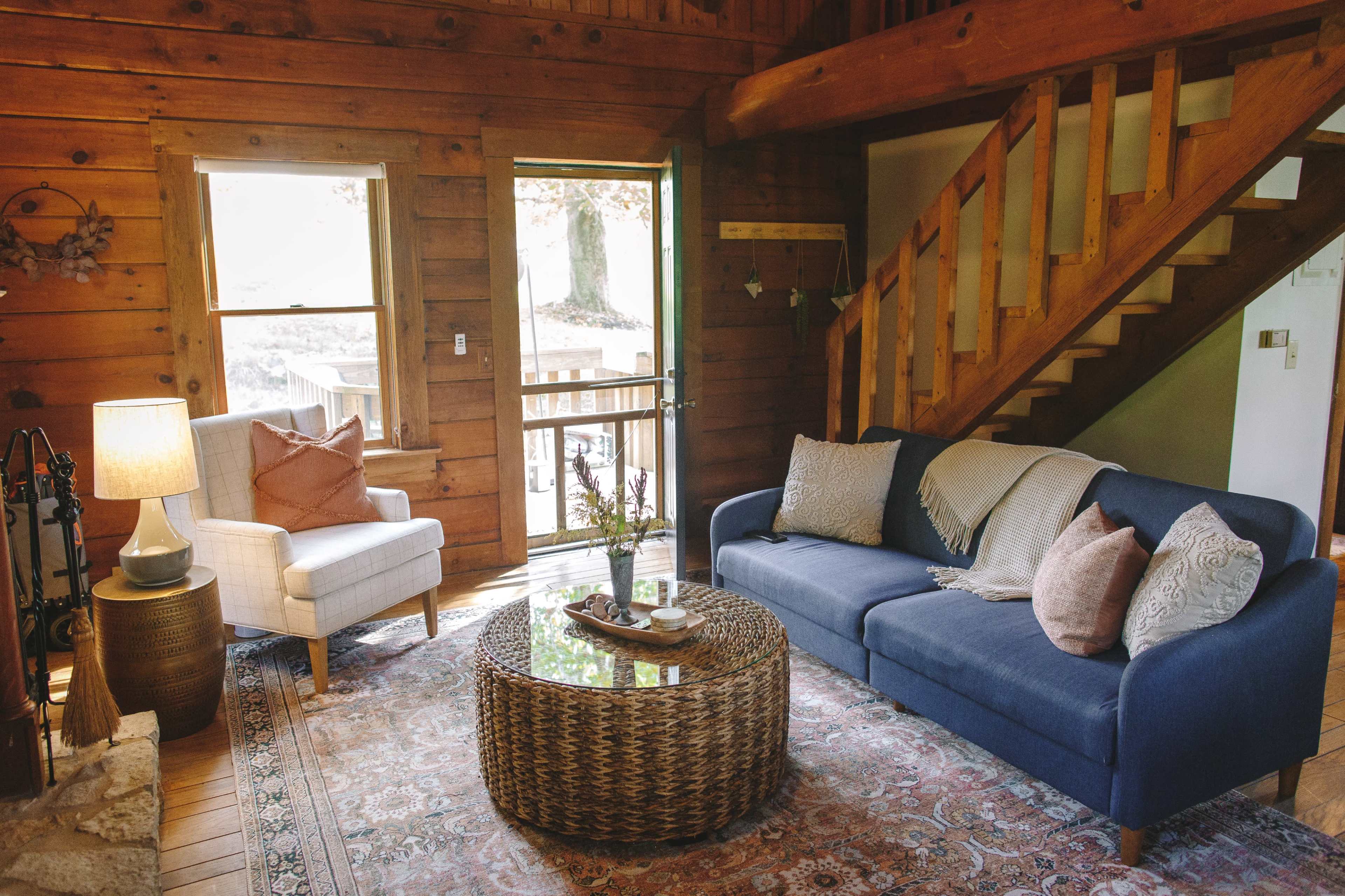 The living room features a blue sofa and a light armchair, a round glass coffee table on a patterned rug, and wooden walls with a staircase in the background.