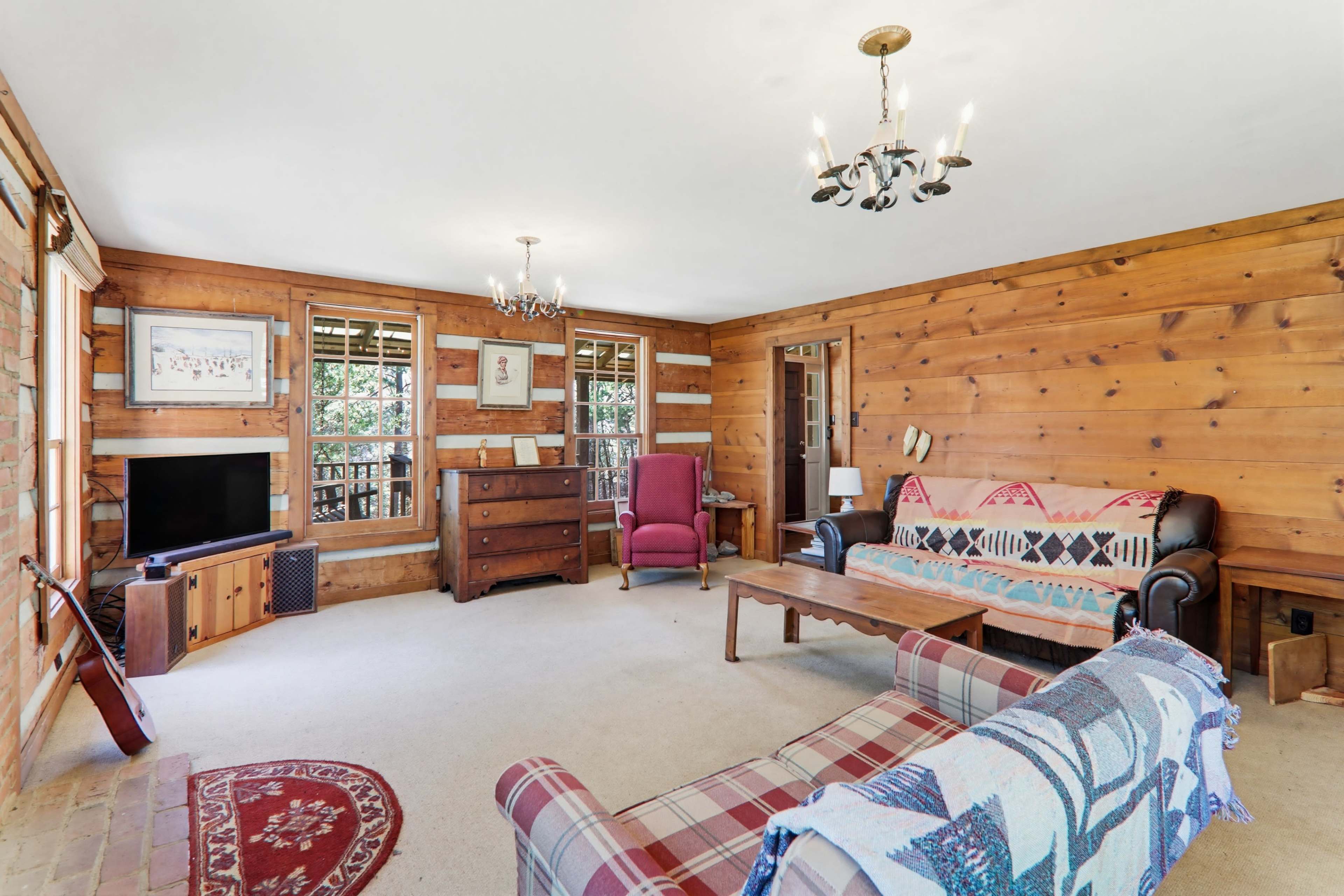 The image shows a cozy living room with wooden paneling, a television, two sofas, a coffee table, and a partial view of the outdoors through large windows.