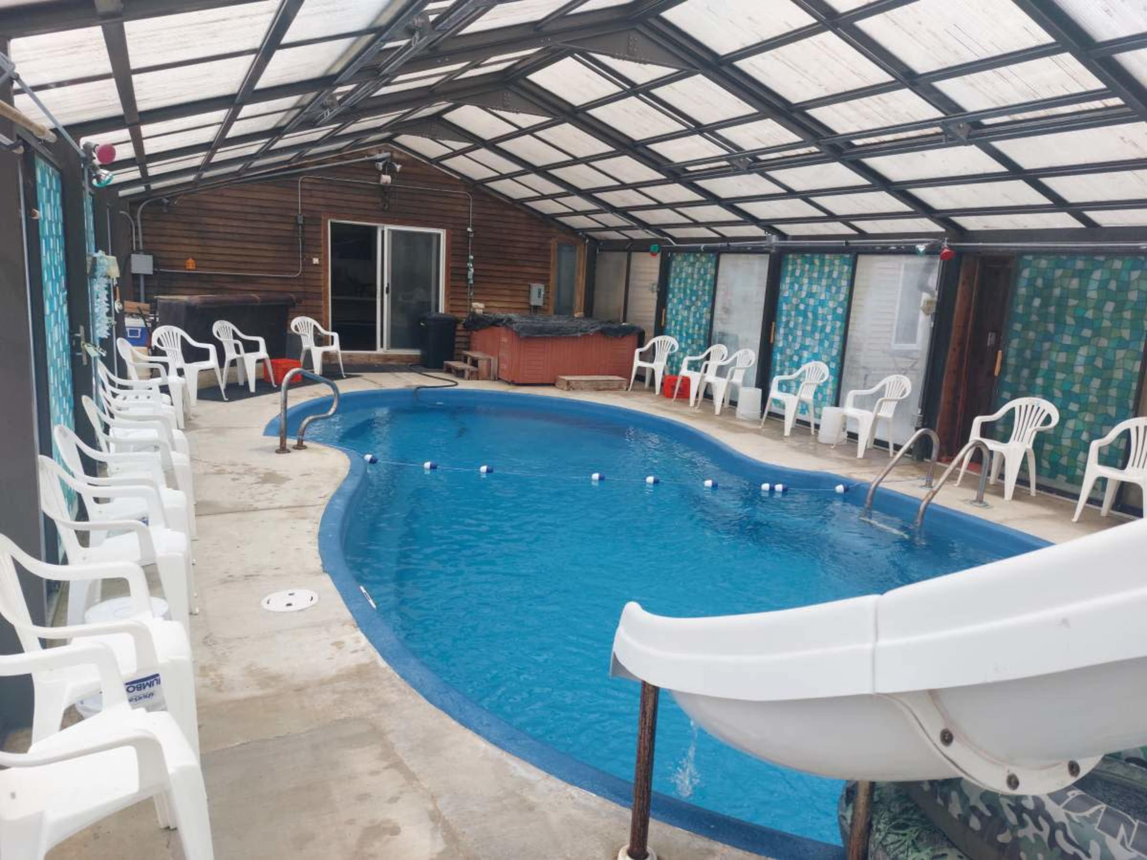 An indoor swimming pool surrounded by white plastic chairs, with a water slide on one side and large windows allowing light to enter.