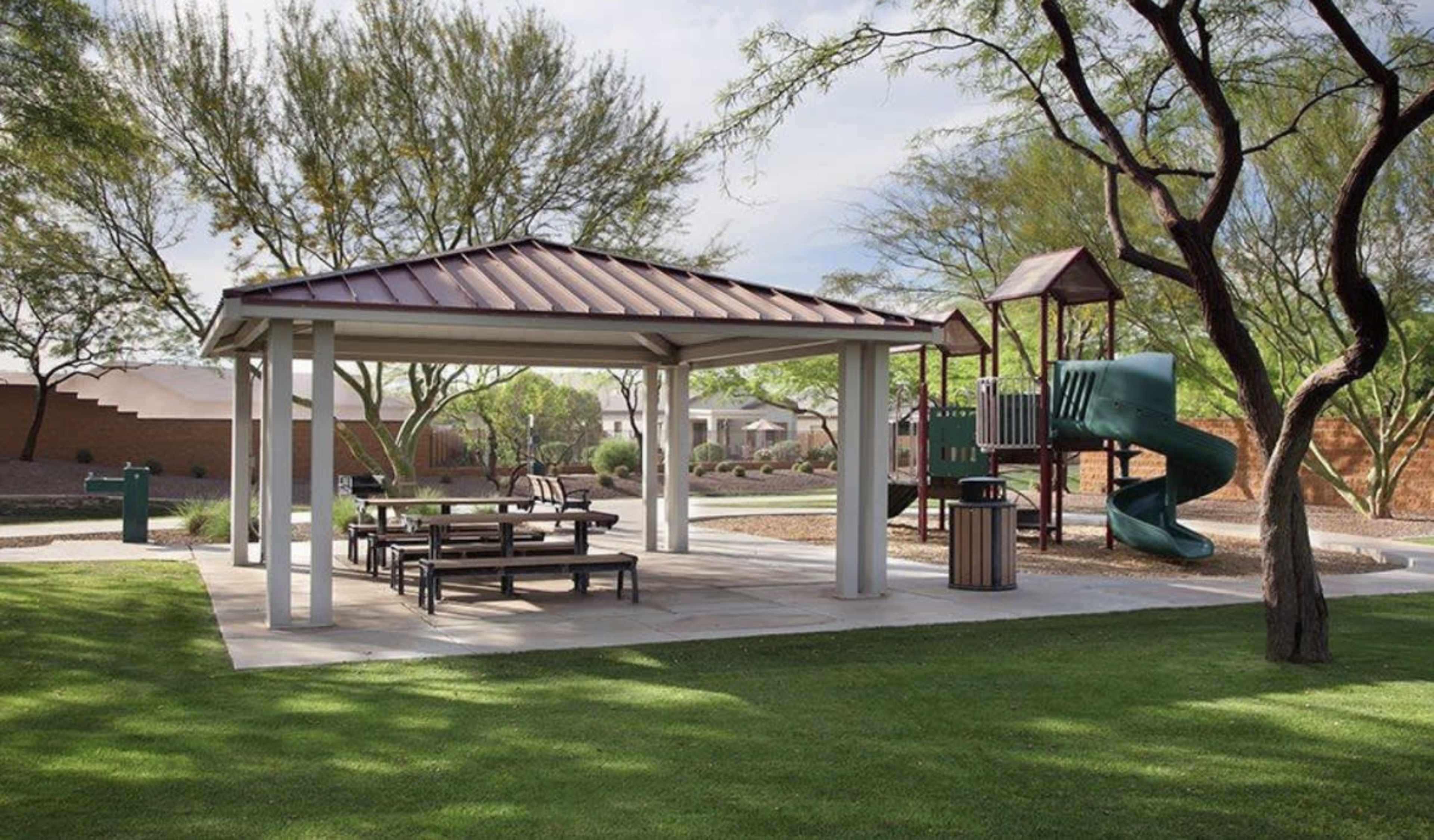 The image shows a covered pavilion with picnic tables next to a playground featuring a slide and climbing structure in a landscaped park area.