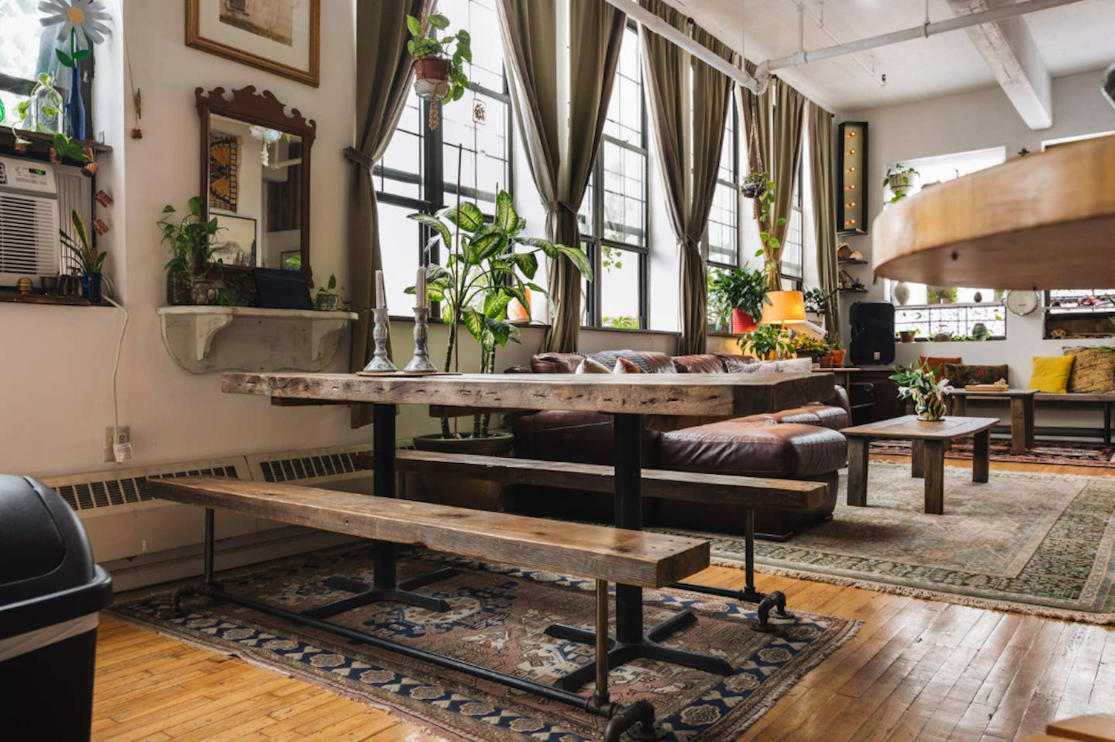 The image shows a cozy living space featuring a wooden dining table with benches, large windows dressed with curtains, and various plants throughout the room.