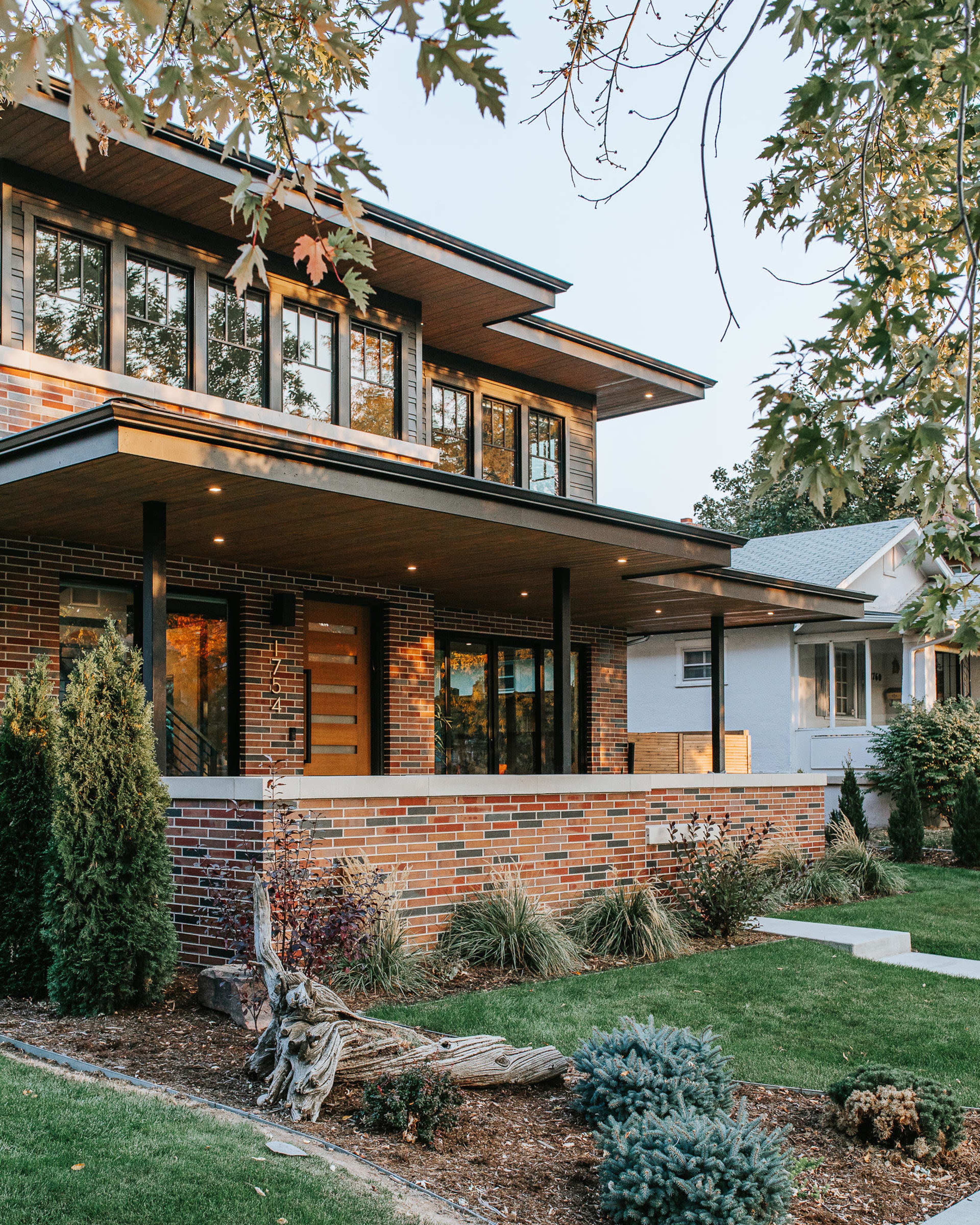 The image shows a modern brick house with multiple windows and a spacious porch, surrounded by landscaped greenery.