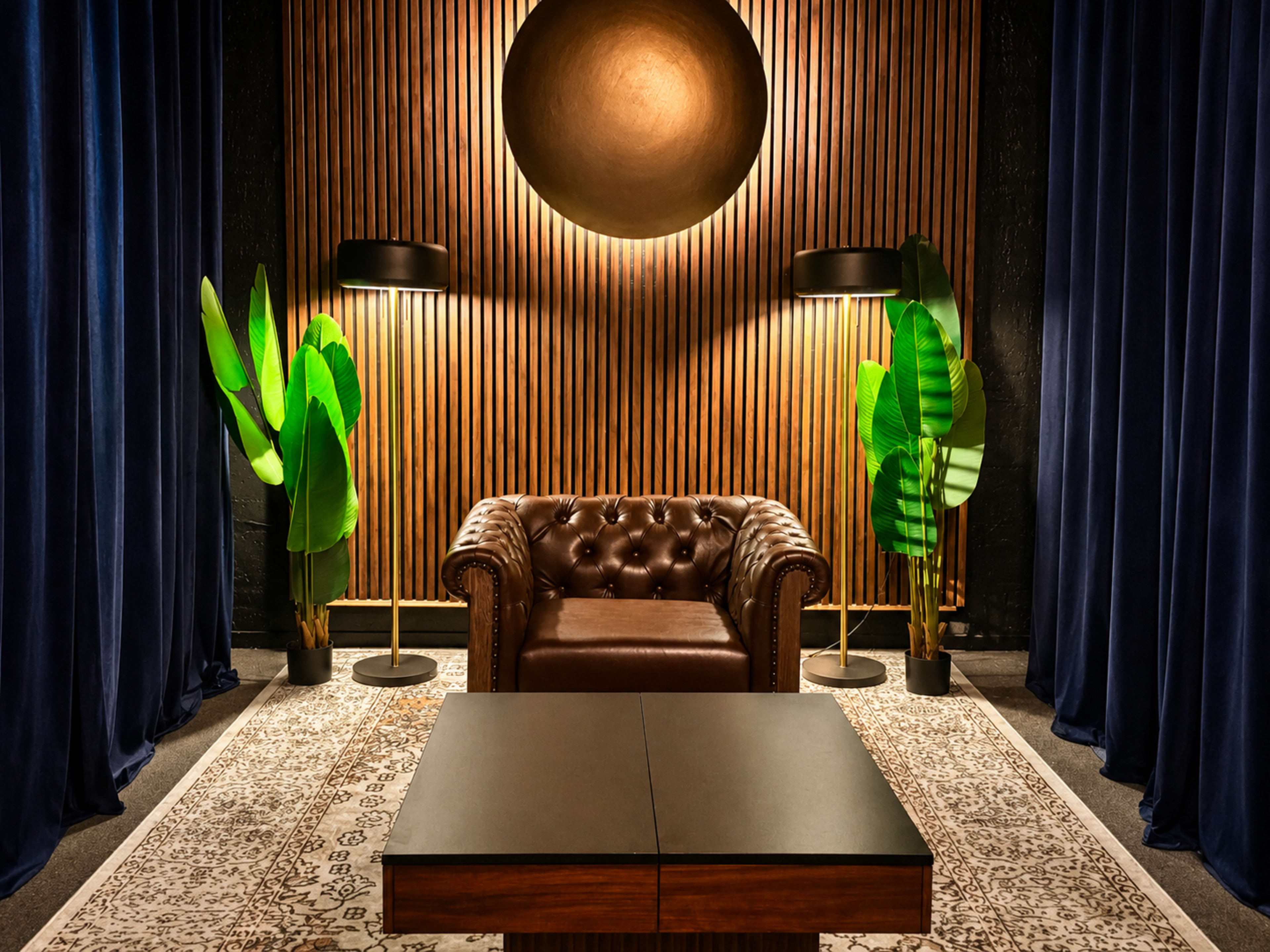 The image shows a cozy lounge area featuring a brown leather Chesterfield sofa, two floor lamps, large green plants, and a decorative round wall piece against a wooden slat backdrop.