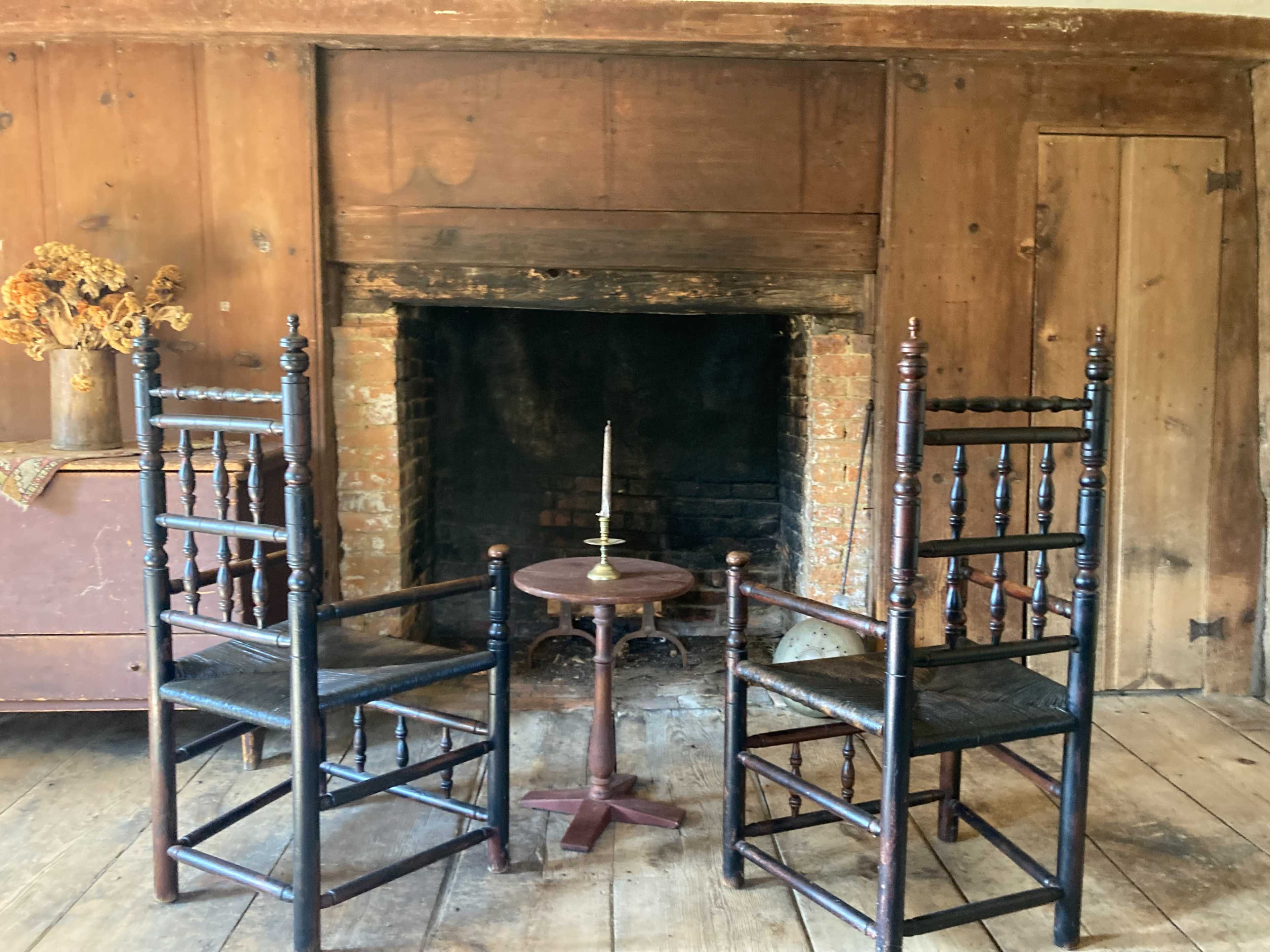 Two wooden chairs face each other across a small table in front of a brick fireplace within a rustic room.